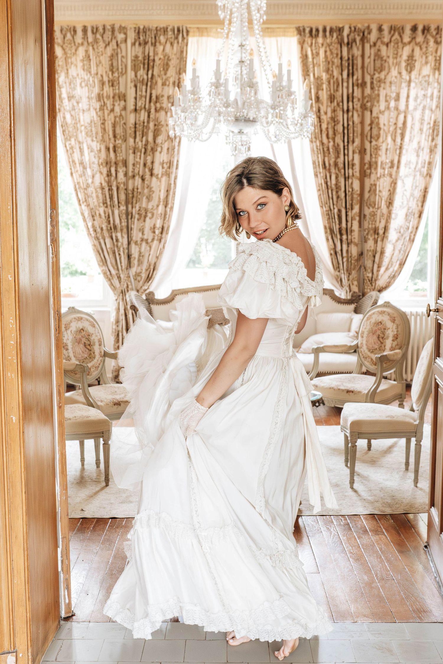 A flowing white dress twirls gracefully in an elegant room with ornate curtains and chandeliers.