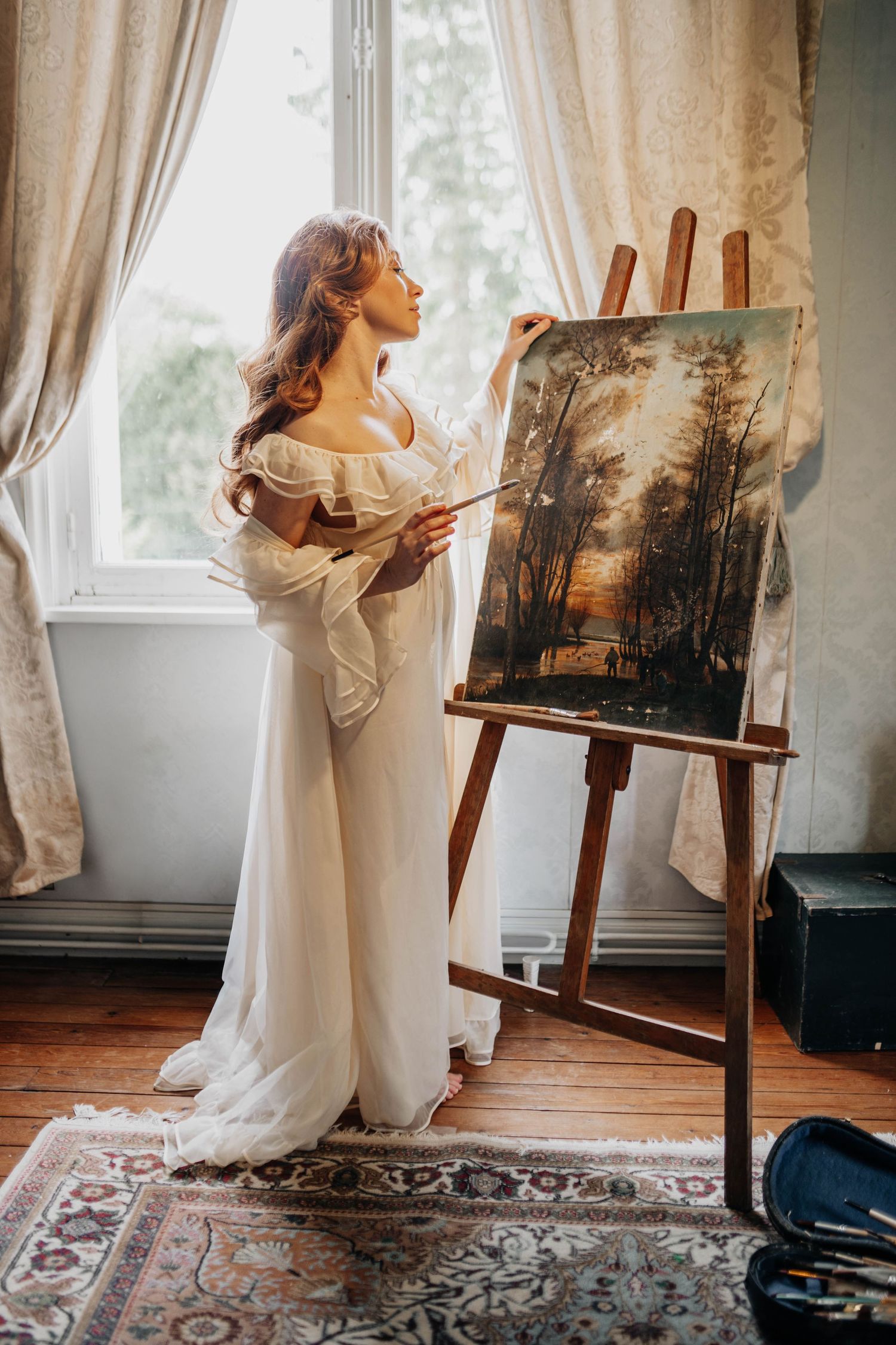 A woman in a white gown painting a forest landscape on an easel by a sunlit window in an elegant room.