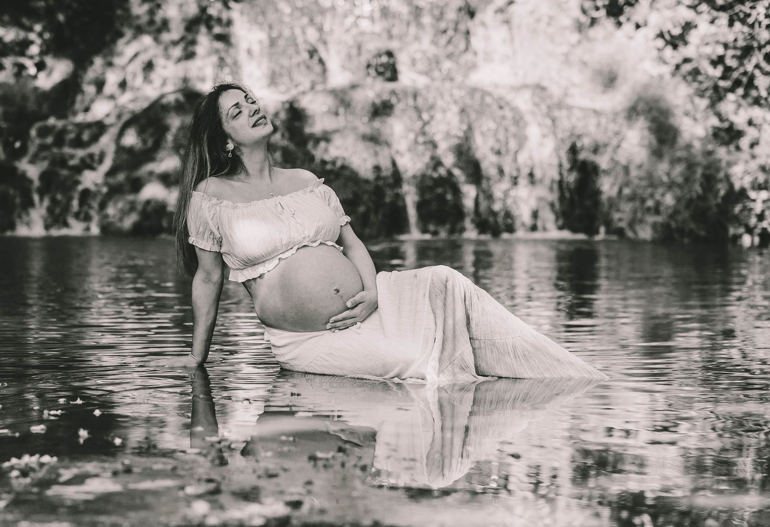 Pregnant woman in white dress poses gracefully in a serene lake, black and white maternity photography.