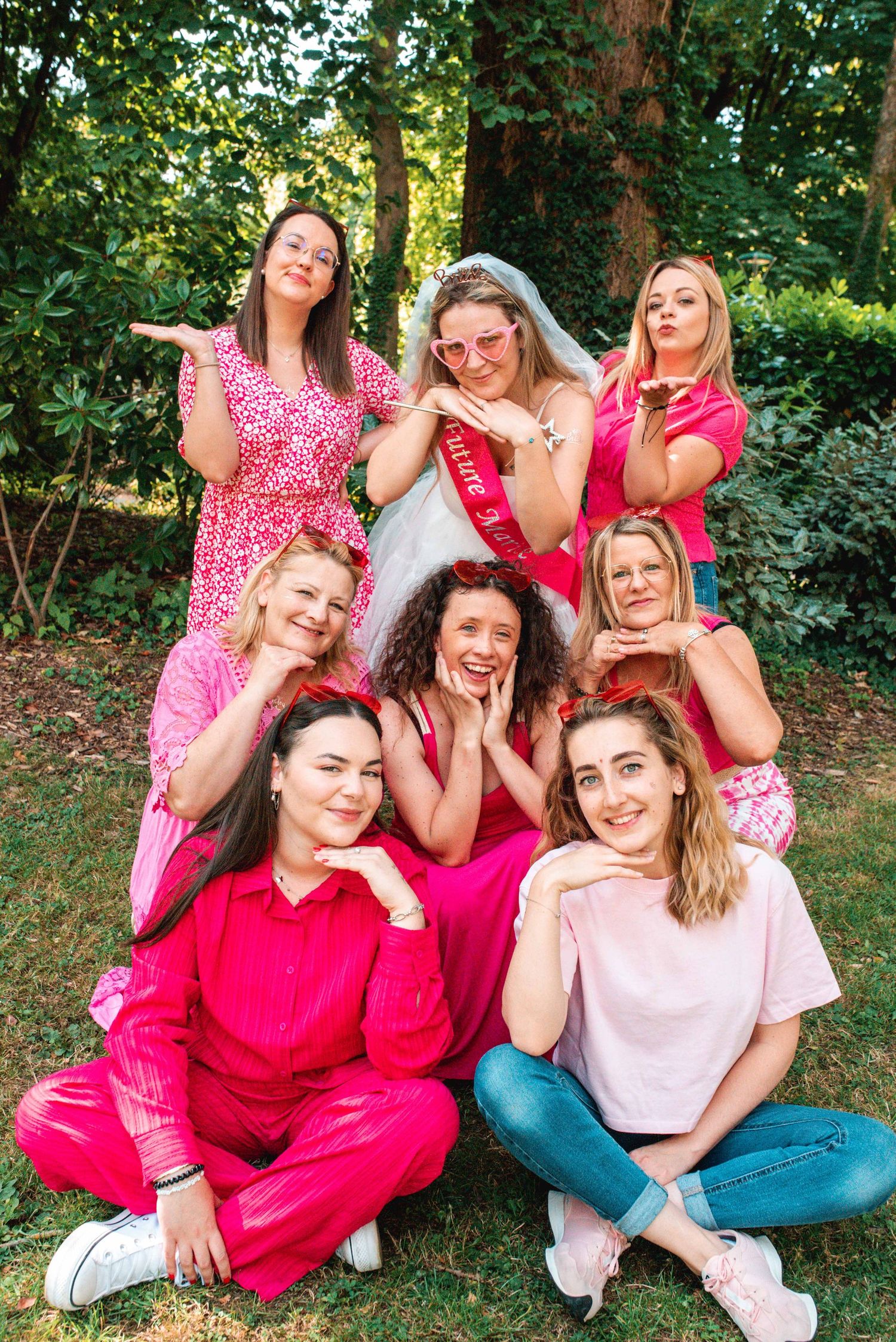 A group in pink and red outfits poses playfully together outdoors in a garden setting.