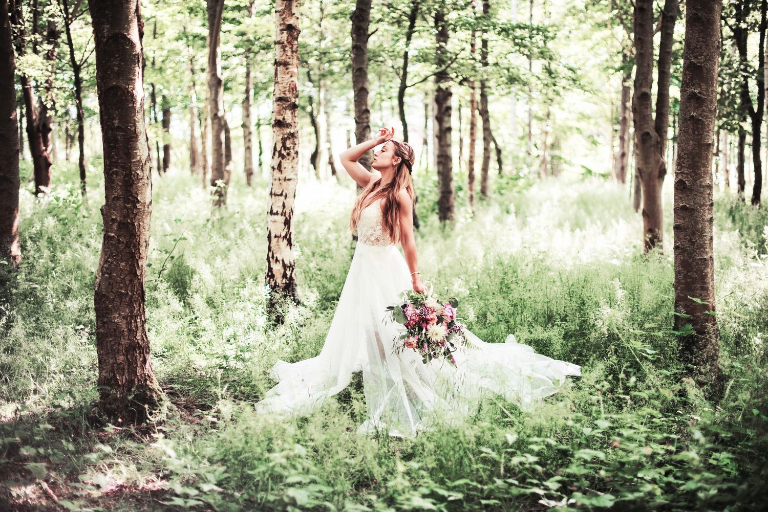 Ethereal wedding dress flows in a dreamy forest setting with pink floral bouquet.