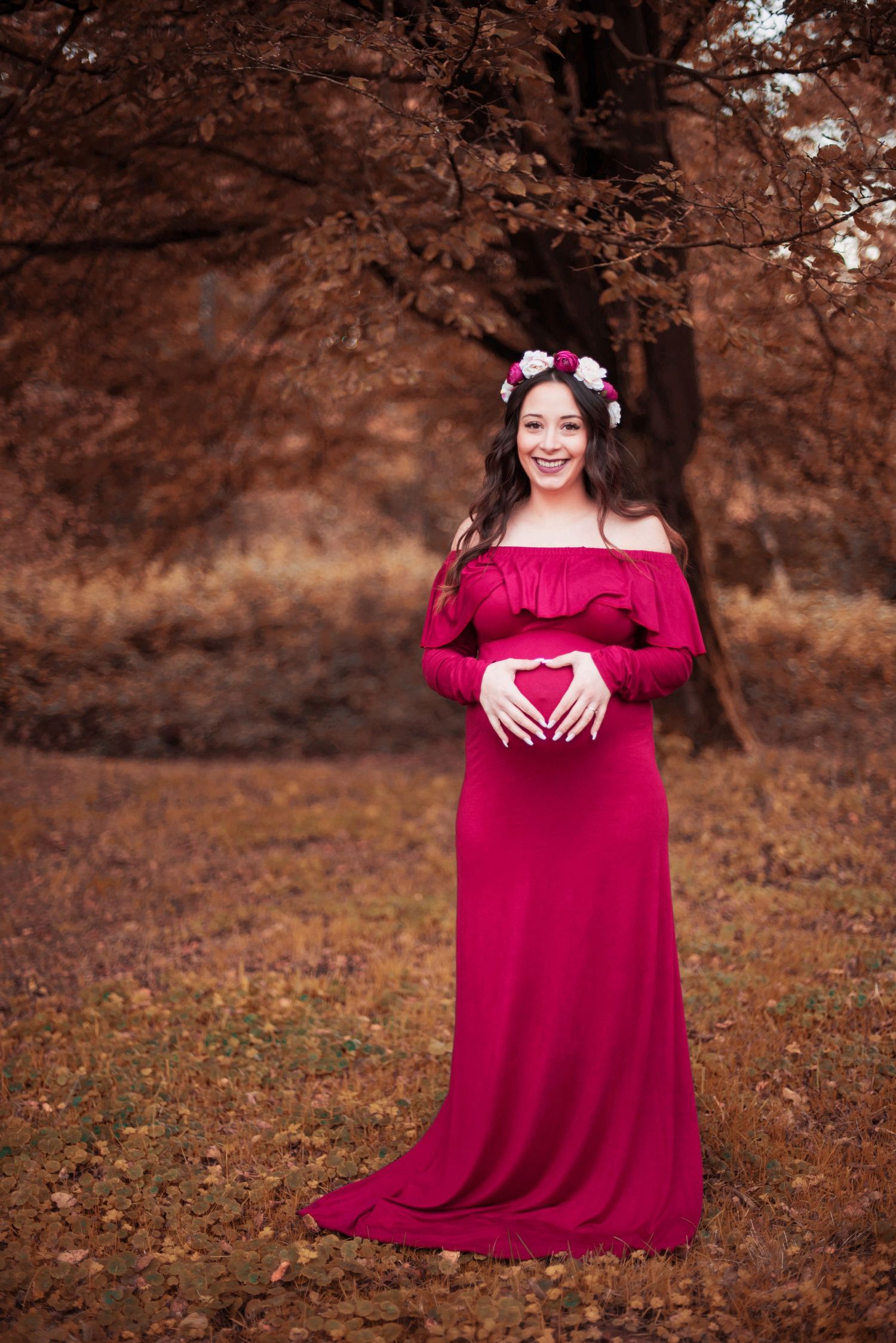 Maternity portrait in flowing burgundy dress with floral crown in autumn setting.