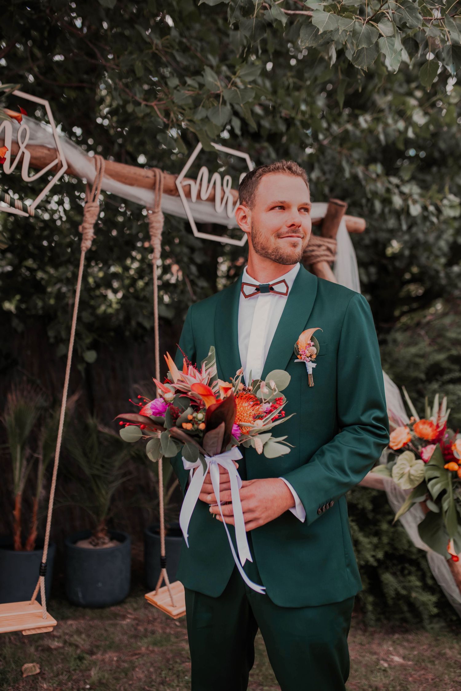 Groom in emerald green suit jacket holding vibrant coral and orange wedding bouquet.