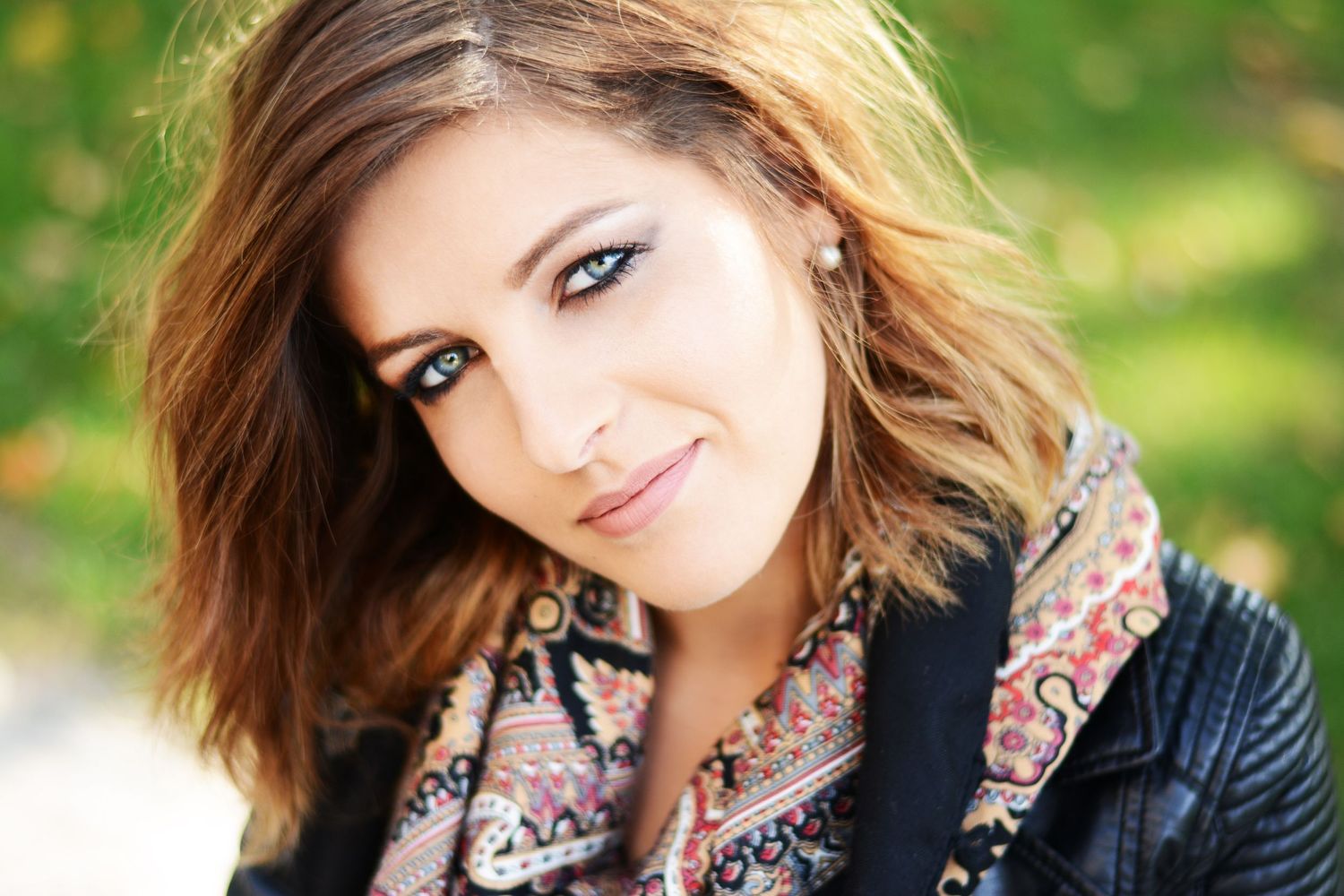 Portrait of person with wavy hair wearing black jacket and patterned scarf outdoors.