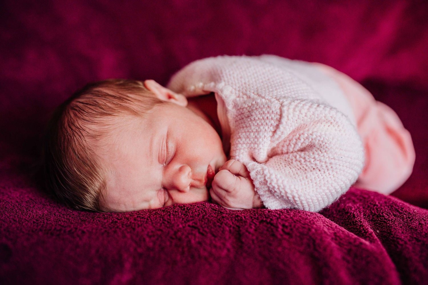 Sleeping newborn baby wearing white knit sweater on burgundy blanket in natural light.