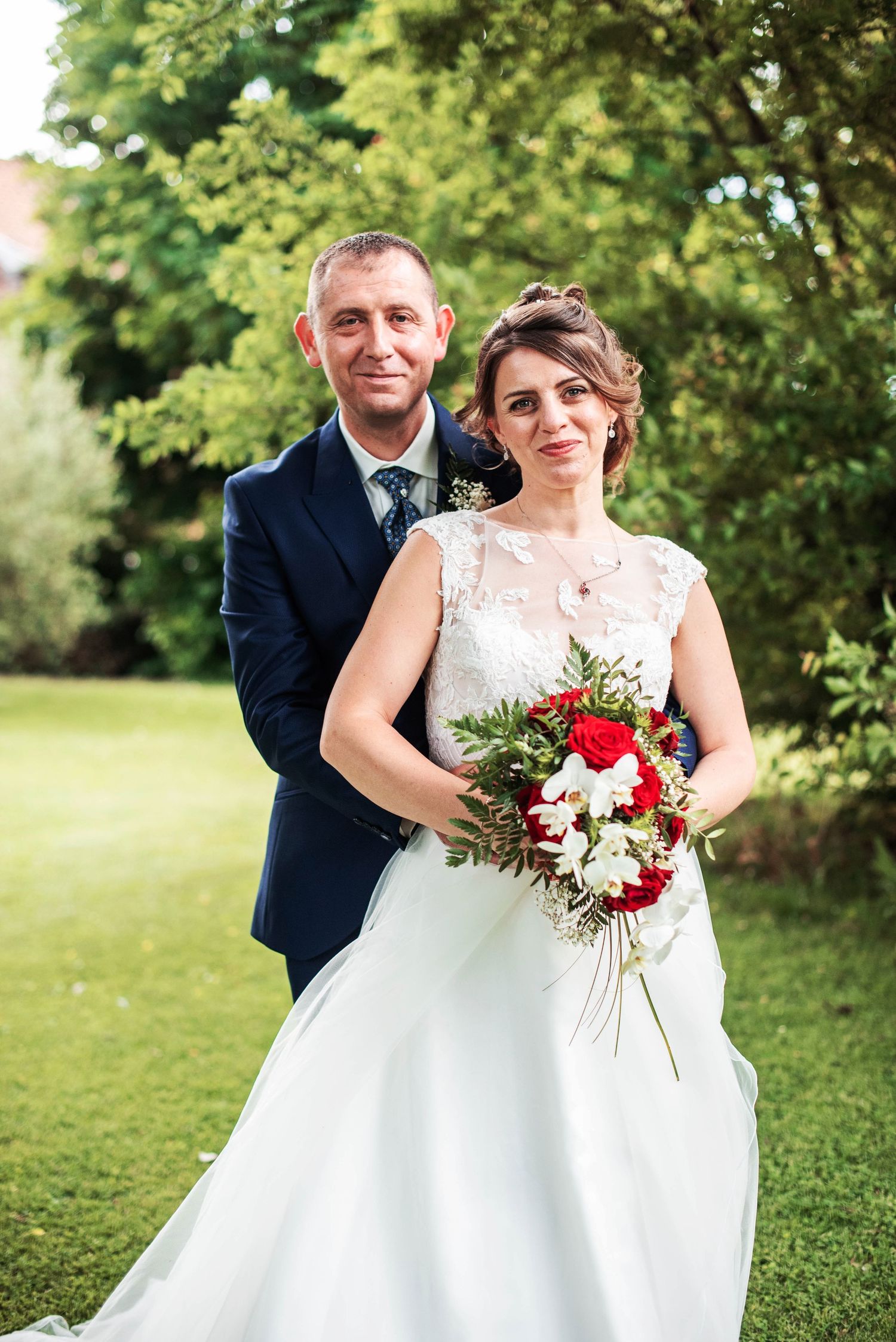 Wedding couple poses in a green garden setting with vibrant red and white bouquet.