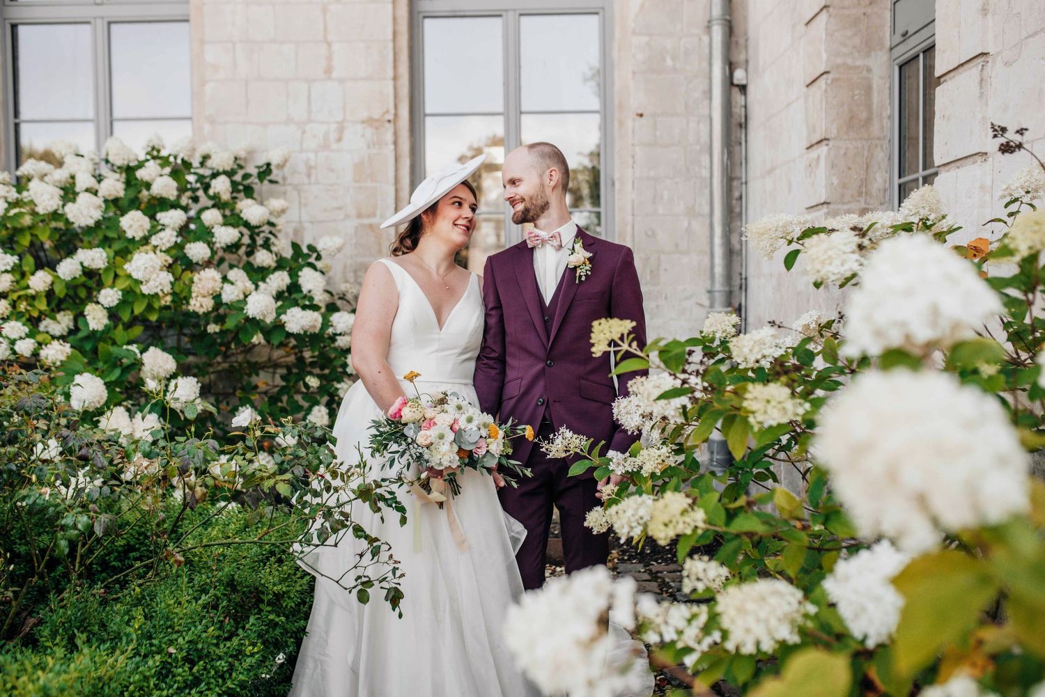 A romantic garden wedding portrait surrounded by blooming white roses and stone architecture.