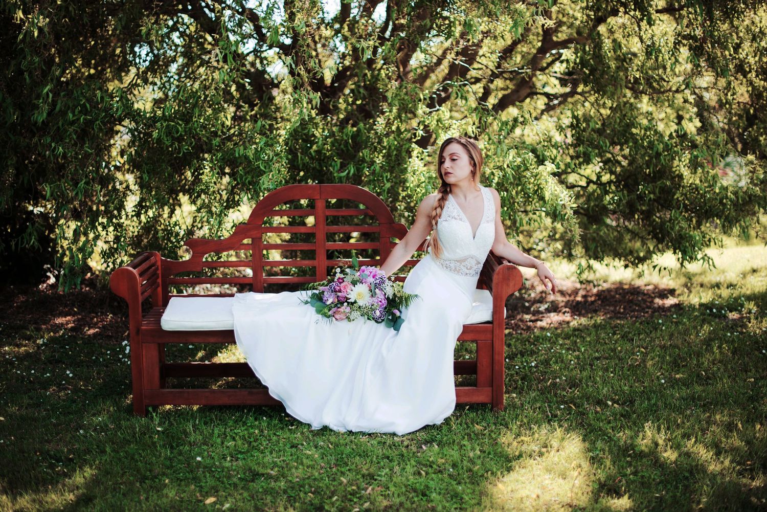 Wedding dress elegantly drapes over red garden bench with vibrant floral bouquet.