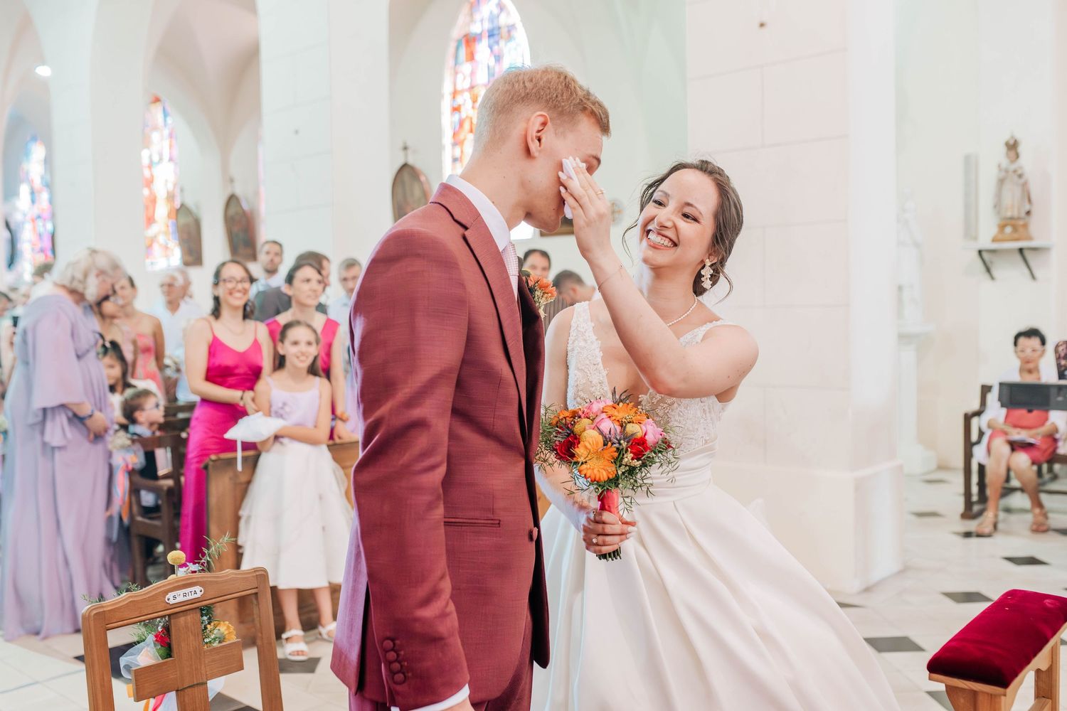 Bride tenderly touches groom's face during church wedding ceremony, holding colorful bouquet in elegant white dress.