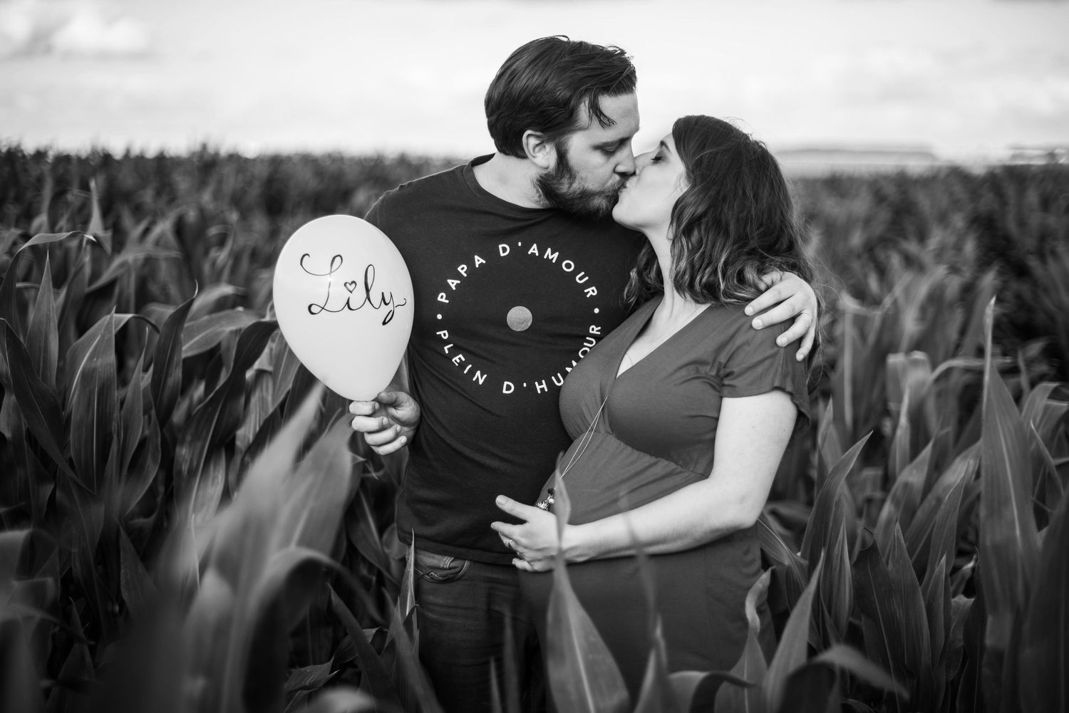 Maternity photo with couple holding balloons in a cornfield, captured in black and white.