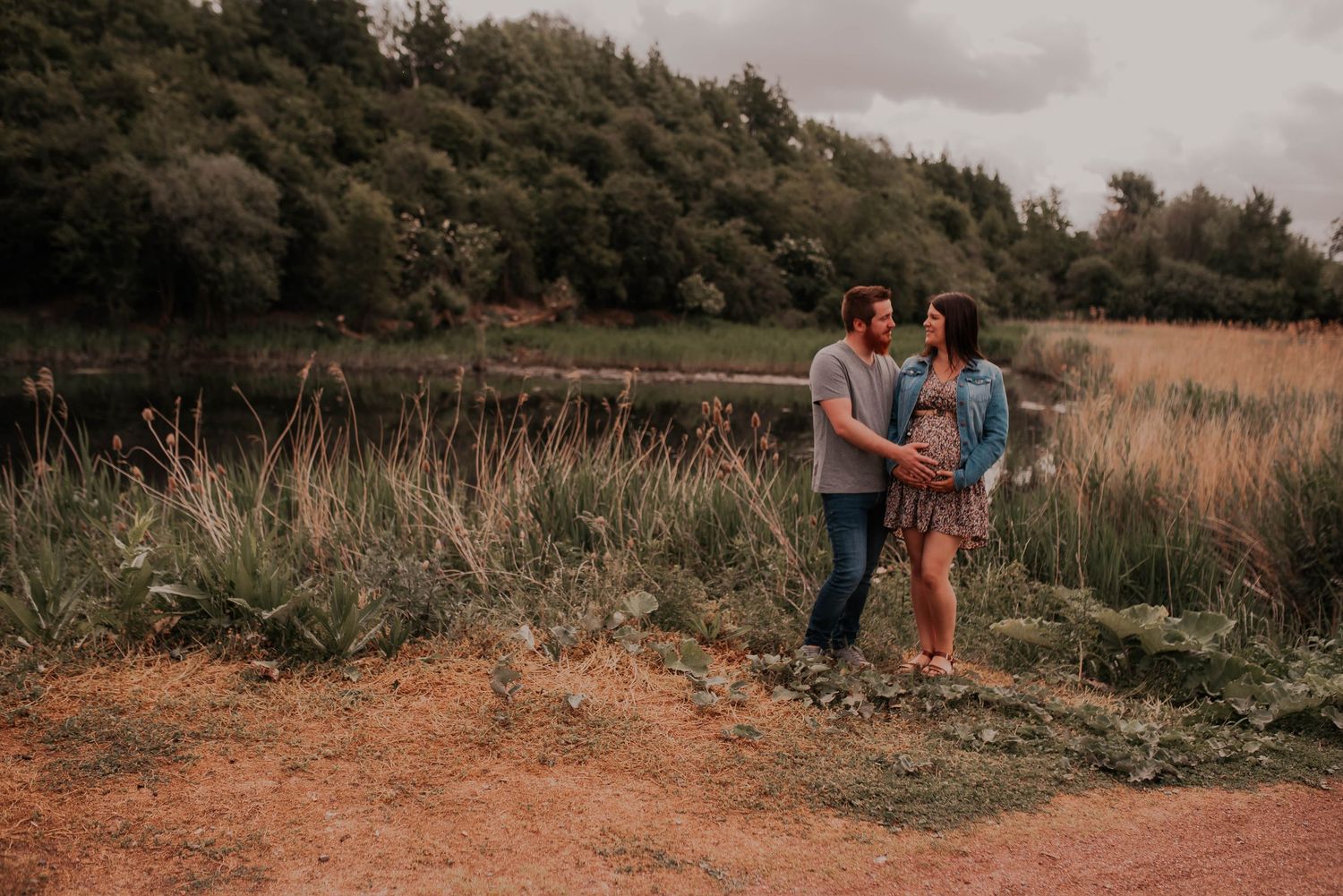 Couple stands together on dirt path with scenic mountain landscape behind them.