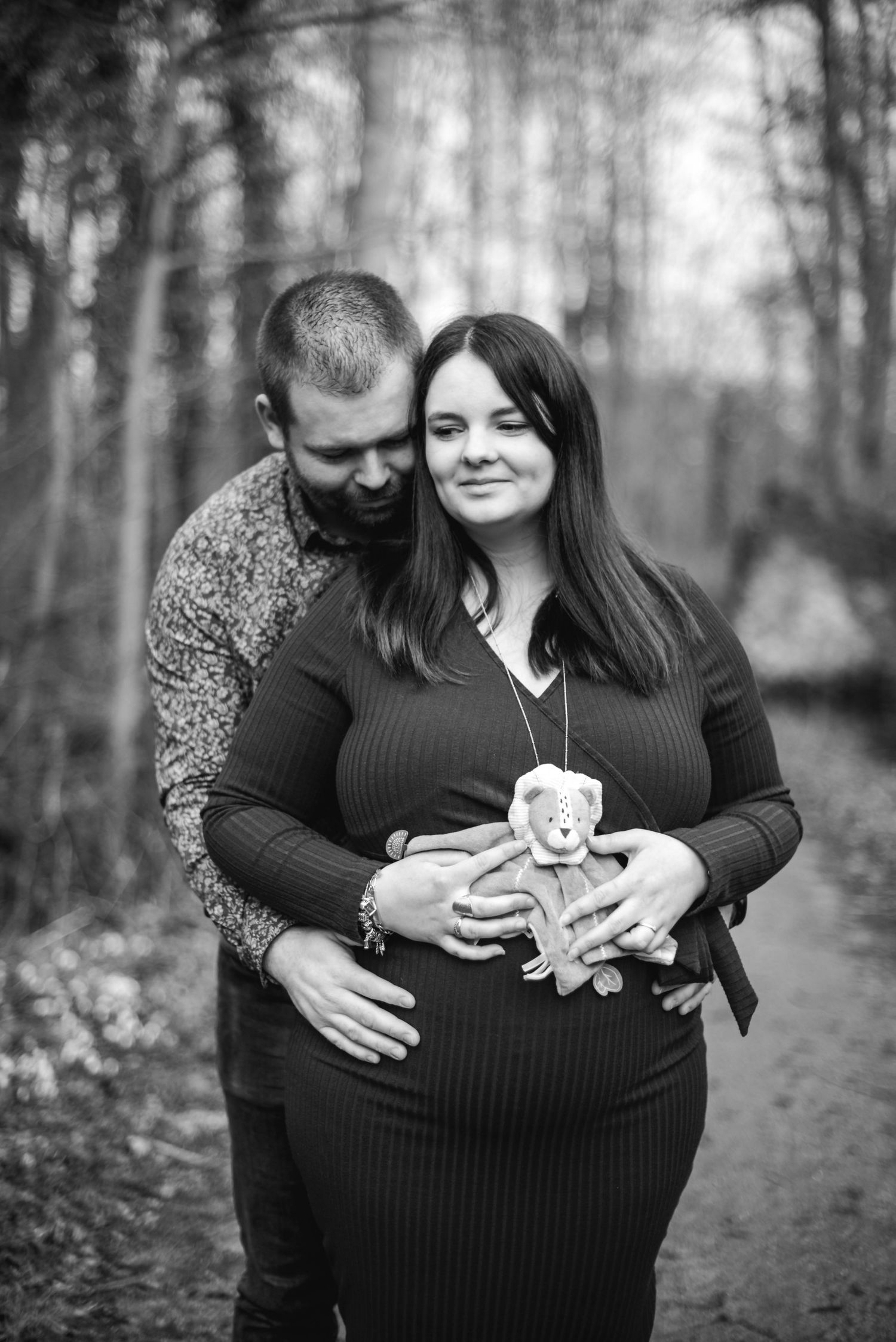 Black and white maternity photo session on a forest path holding baby items.