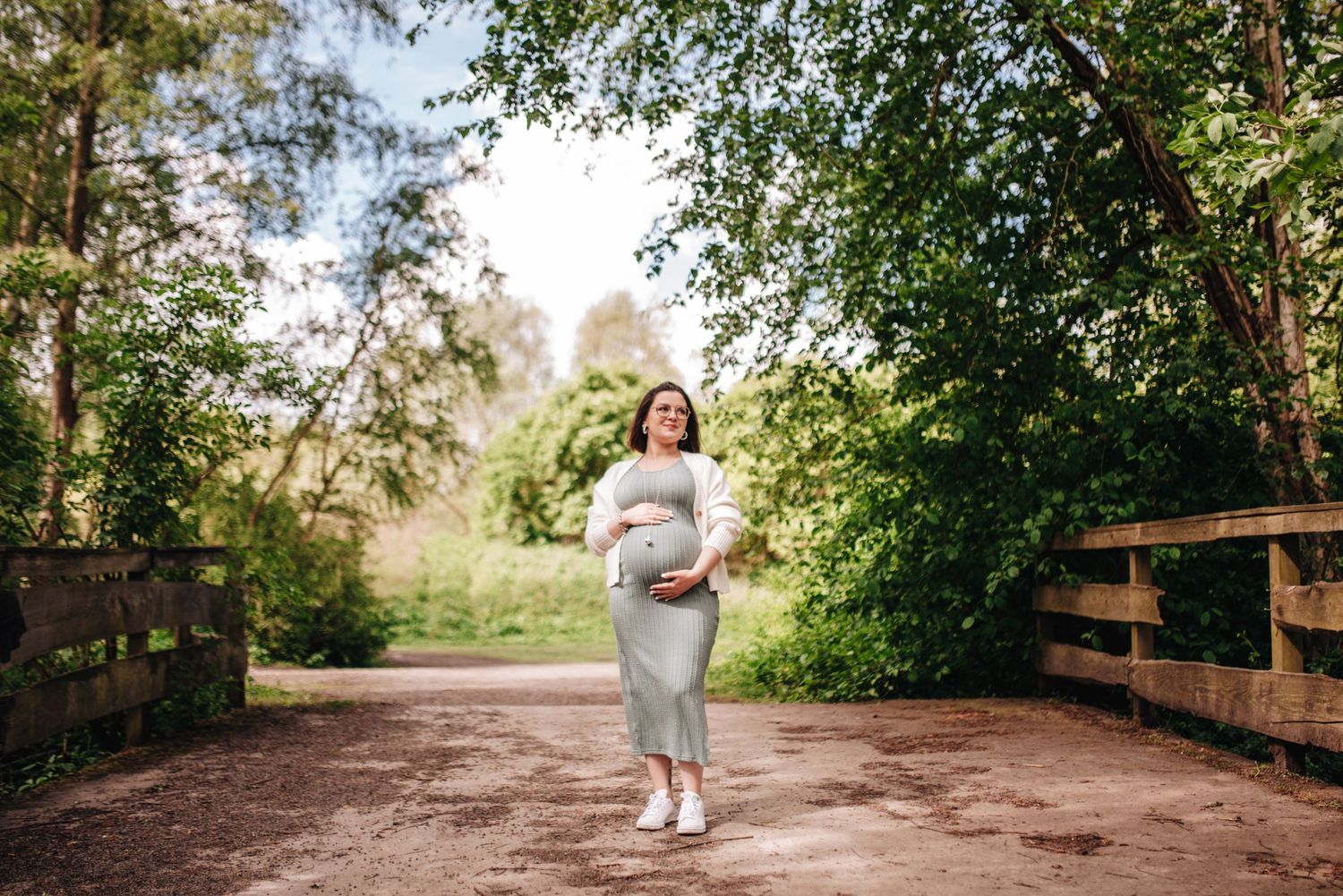 A pregnant woman in a grey dress walking on a wooden bridge path surrounded by greenery.