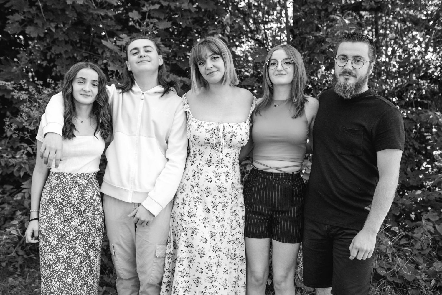 A black and white portrait of five people standing together against a leafy background.