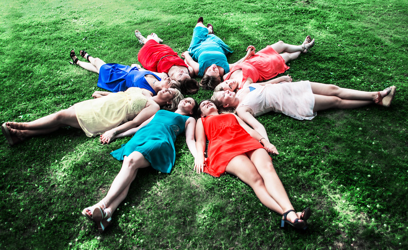 Friends form a colorful star shape lying on green grass wearing bright dresses.