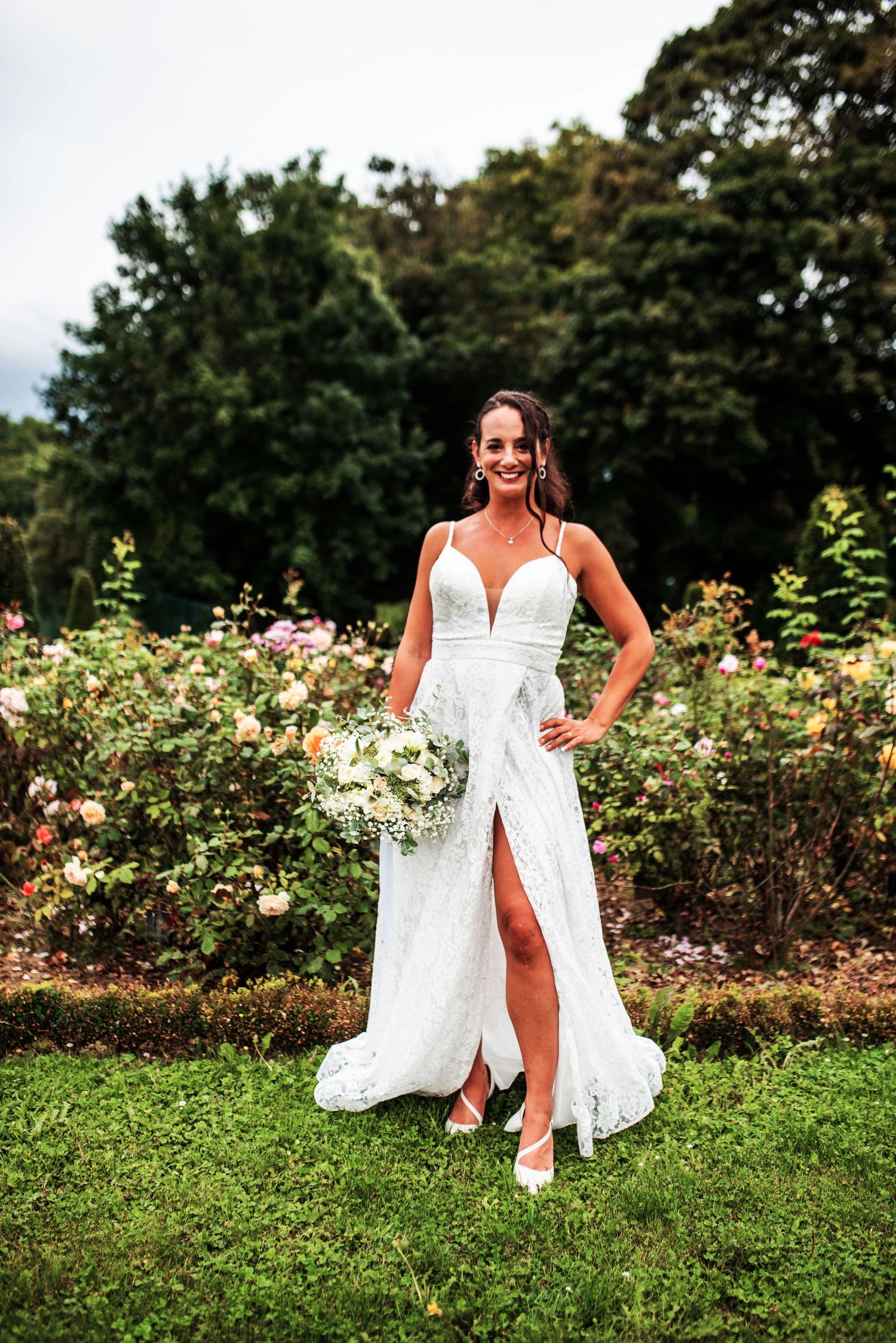 Bride poses playfully in a white wedding dress near blooming rose bushes in a garden.