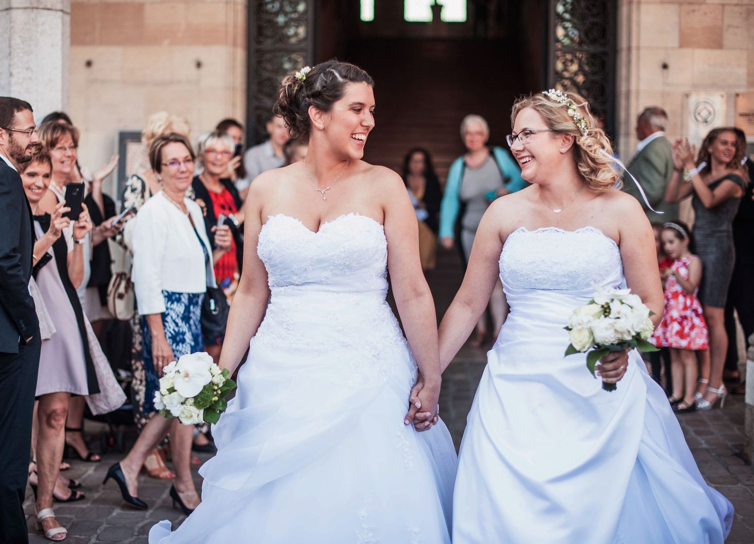 Two brides in matching white dresses exit their ceremony while guests look on with joy.