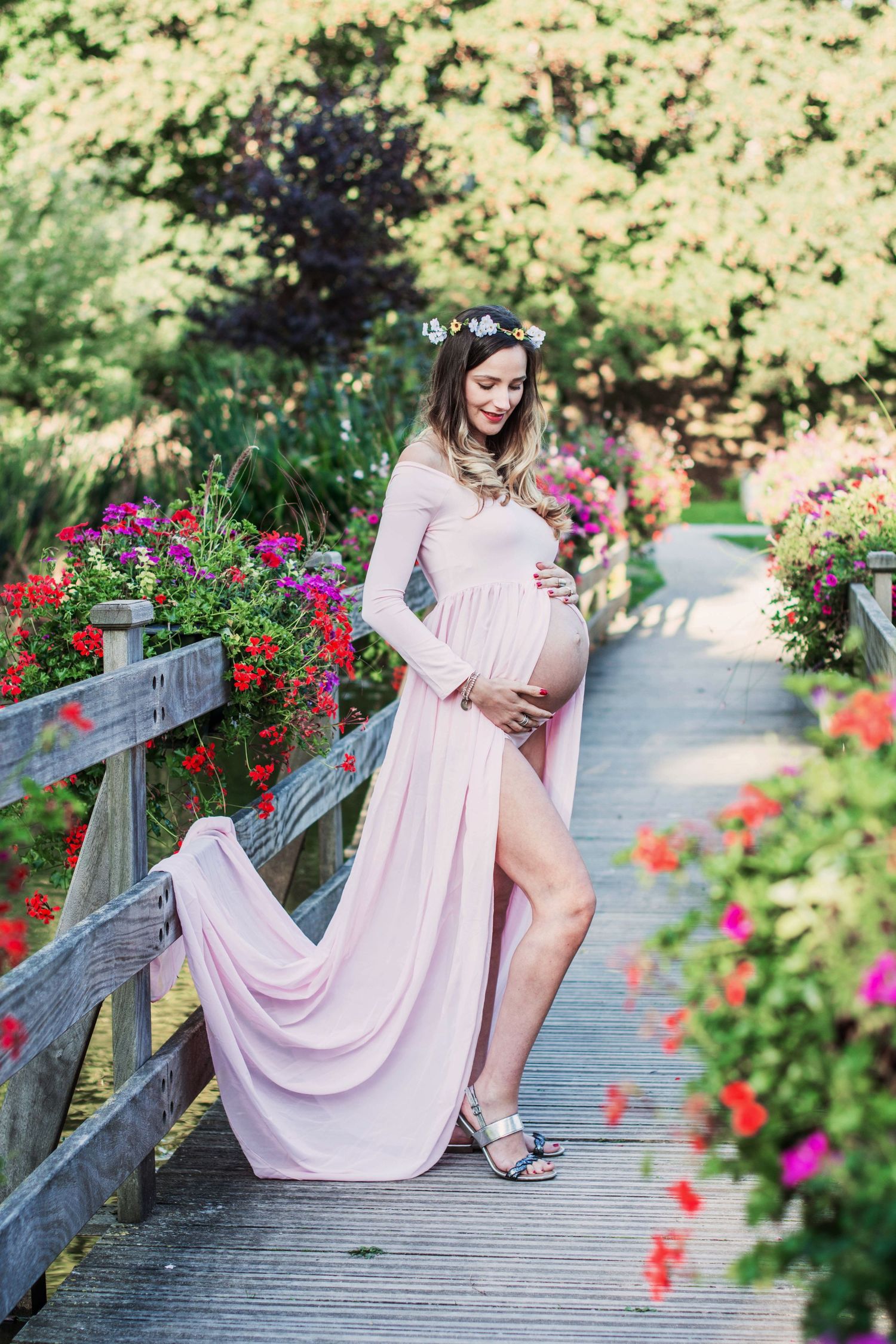 Maternity photos on wooden bridge surrounded by vibrant red flowers and greenery.