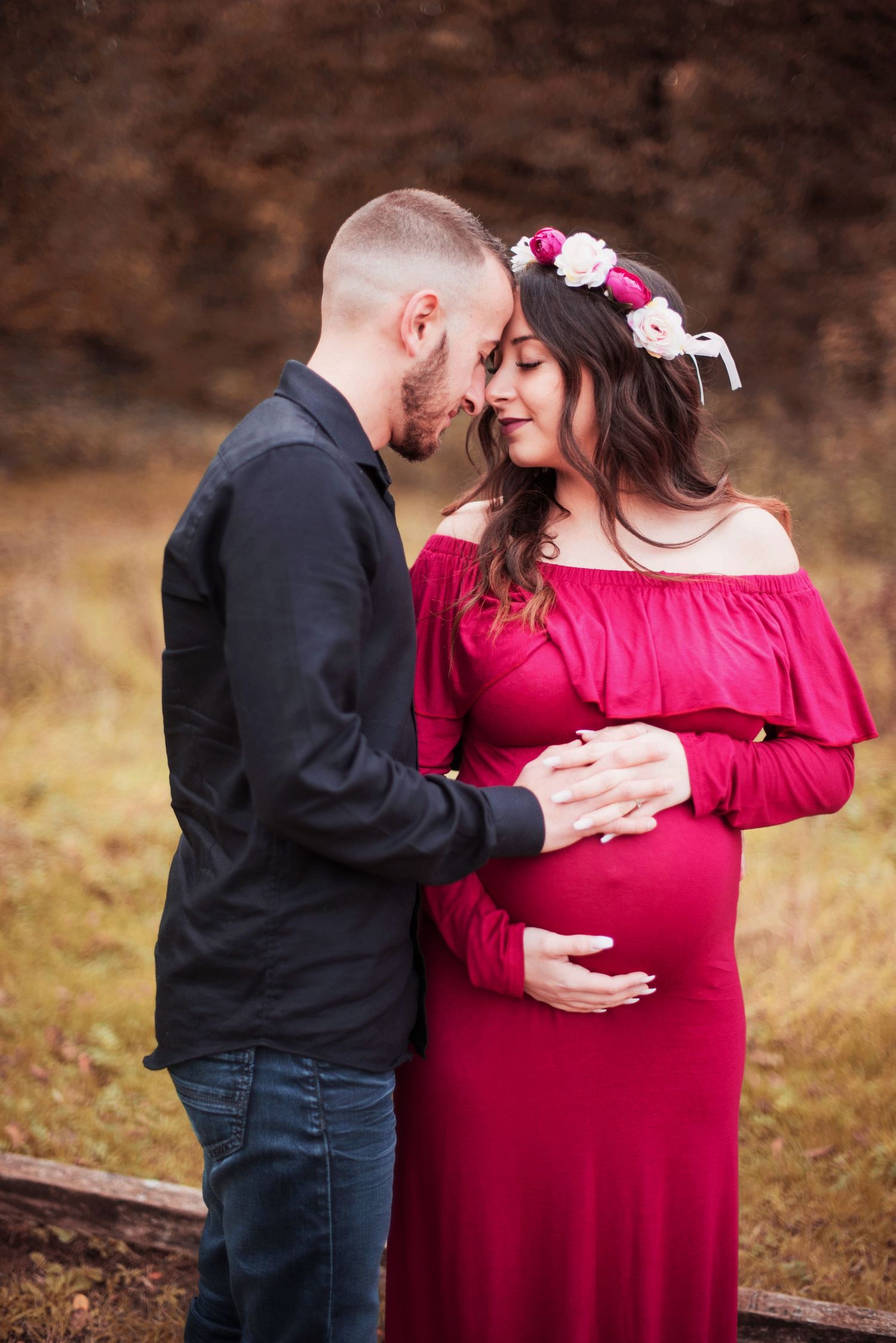 A romantic maternity photo showing a couple in a field with the woman wearing a red off-shoulder dress and floral crown.