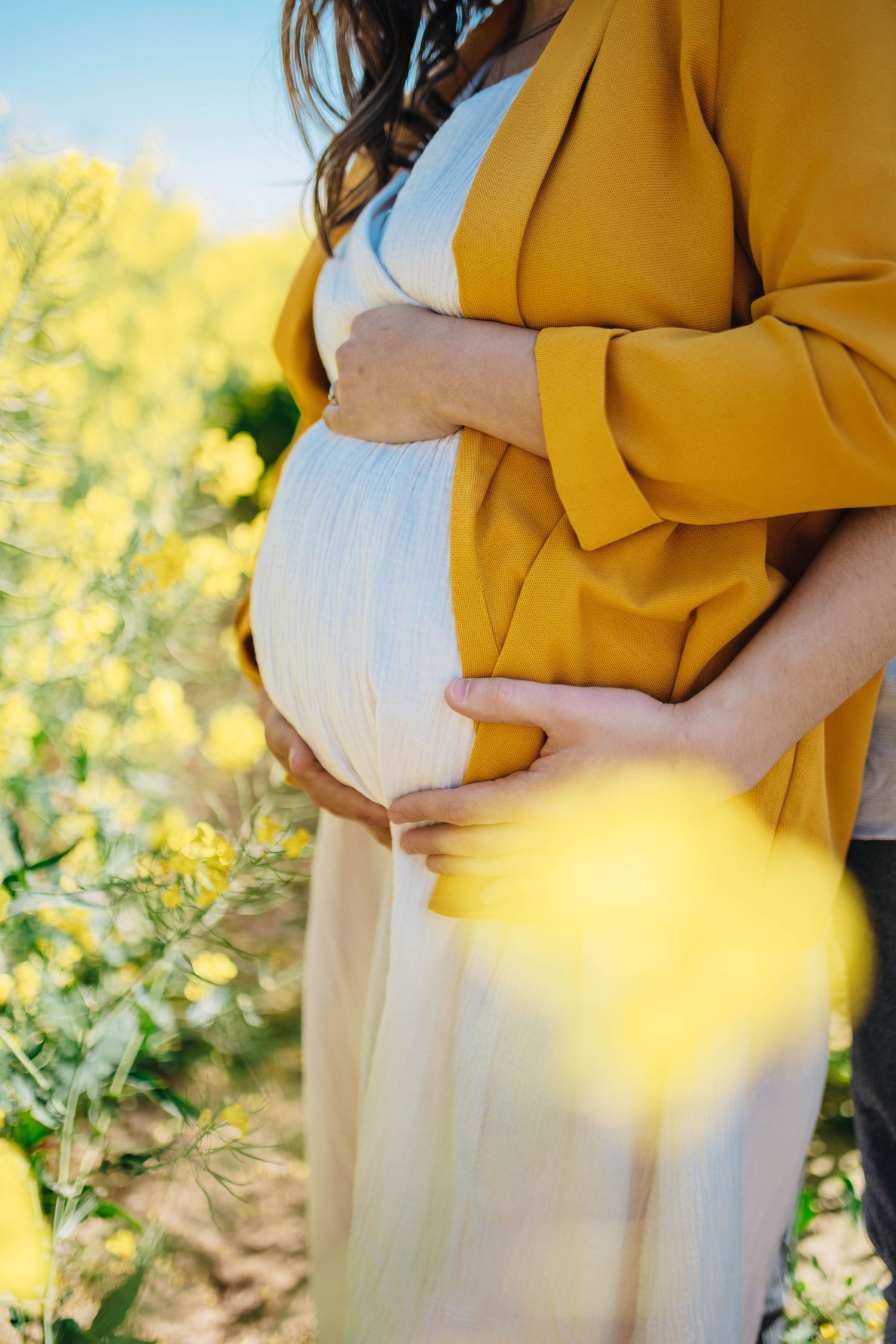 Pregnant woman in white dress and yellow jacket cradling baby bump in a vibrant yellow flower field.