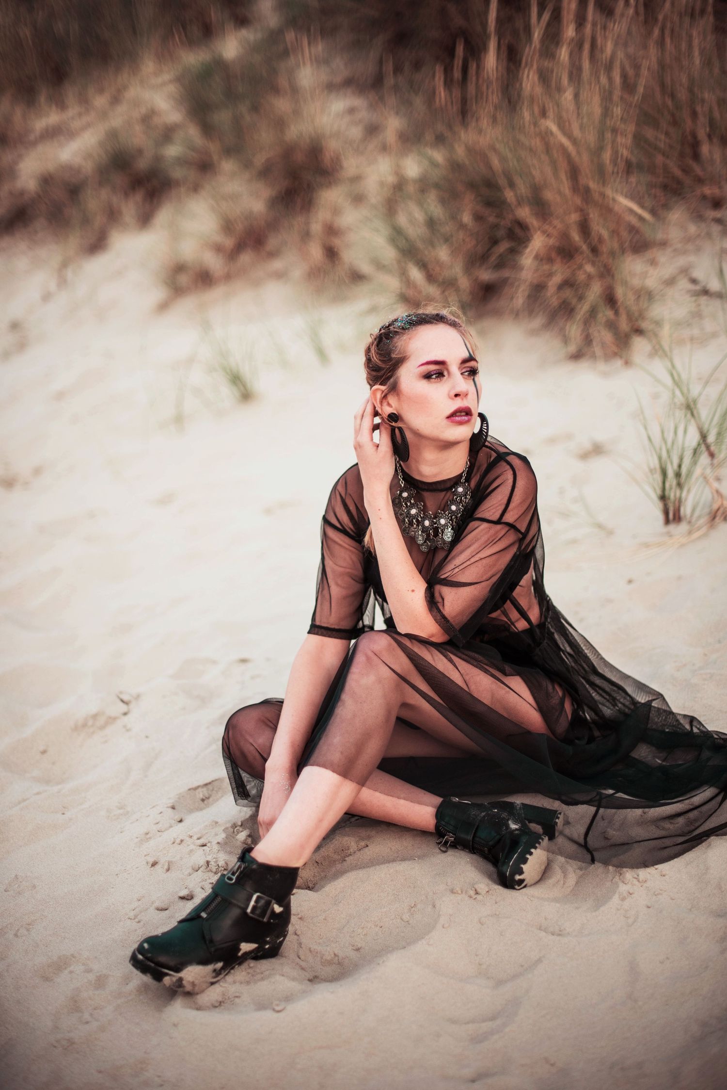 Figure in flowing black dress reclines gracefully on sandy beach during outdoor photoshoot.