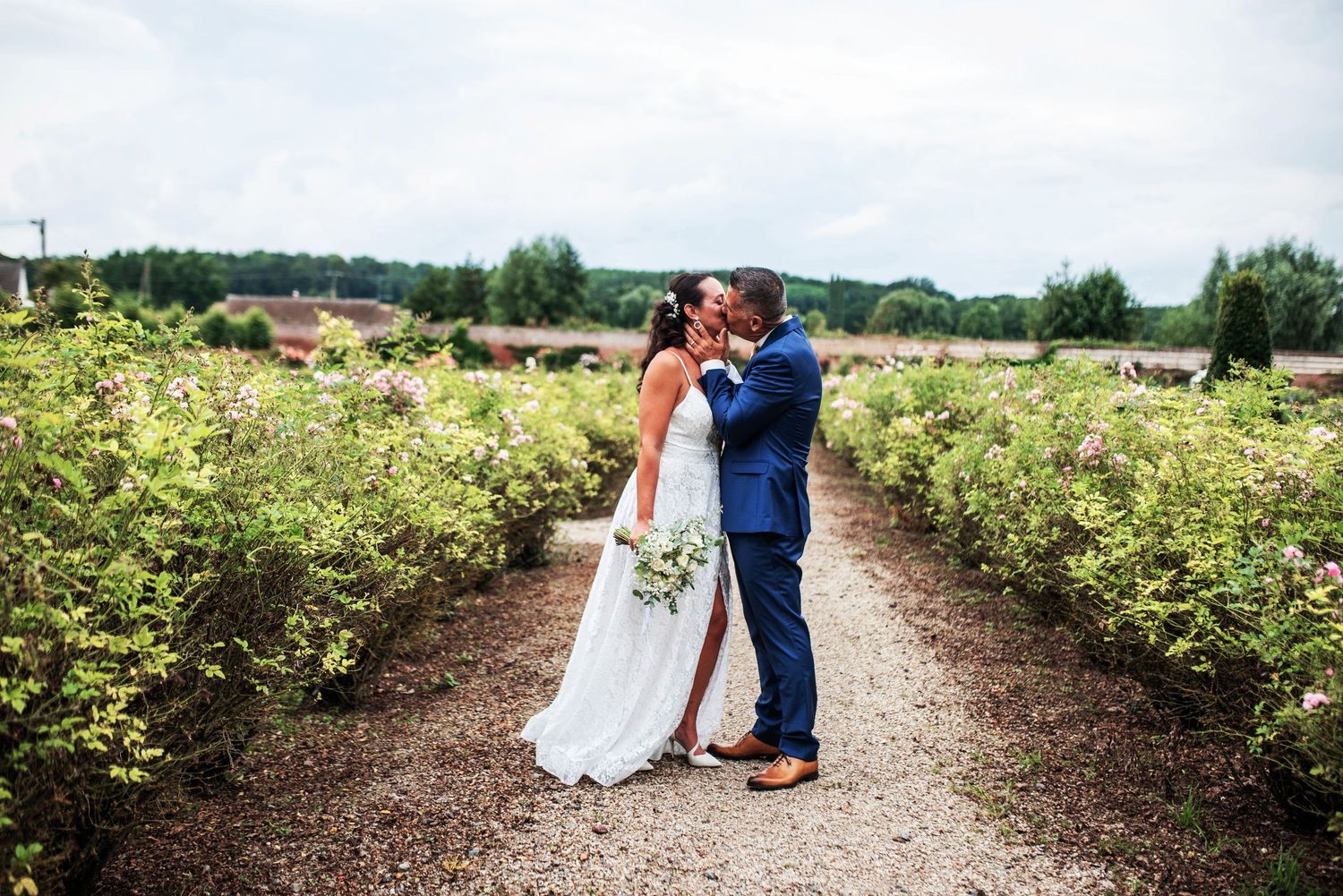 Newlyweds stroll along a garden path surrounded by flowering bushes.
