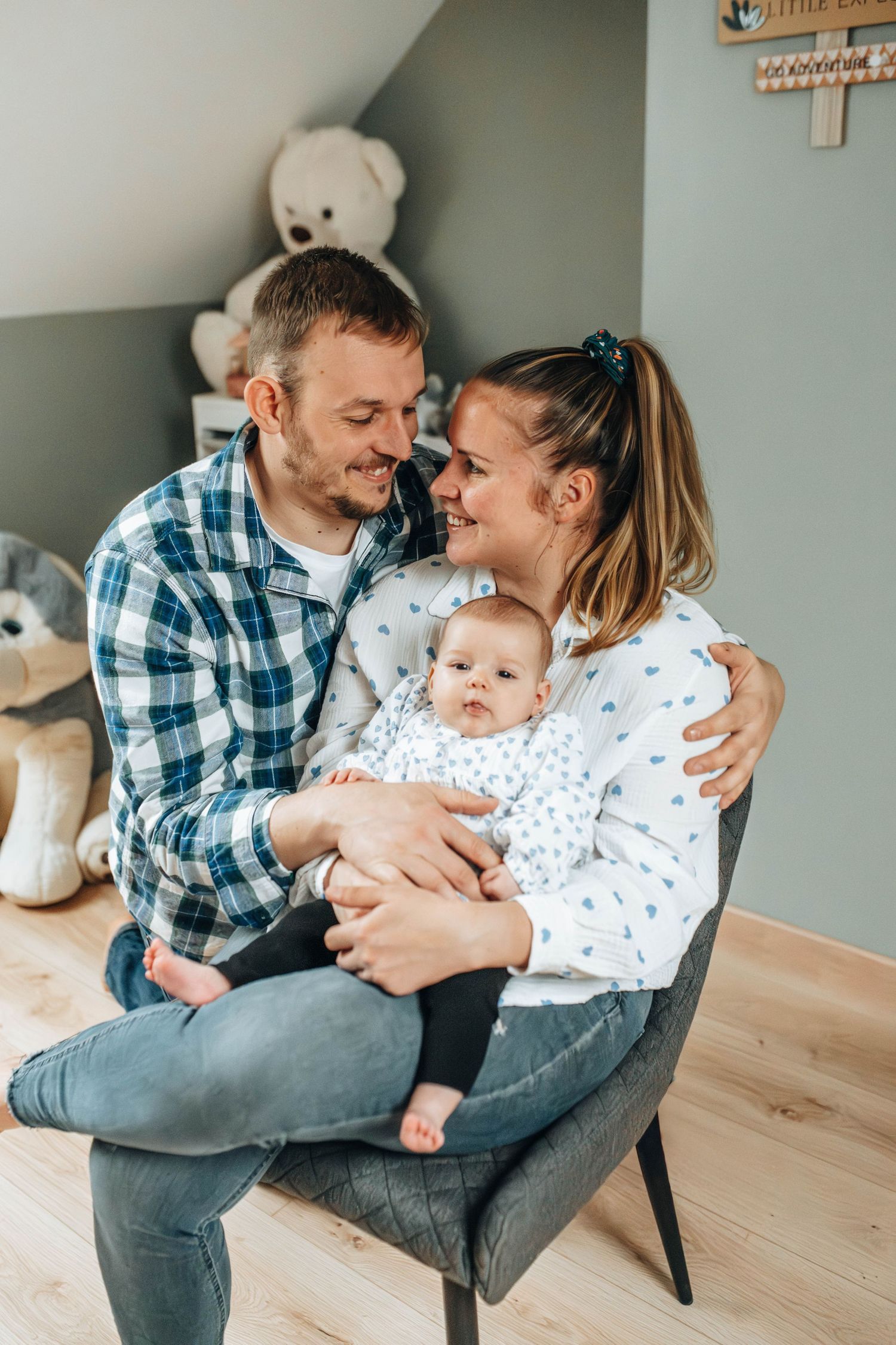 Family with baby sitting together on grey chair in living room with plush toys visible in background.
