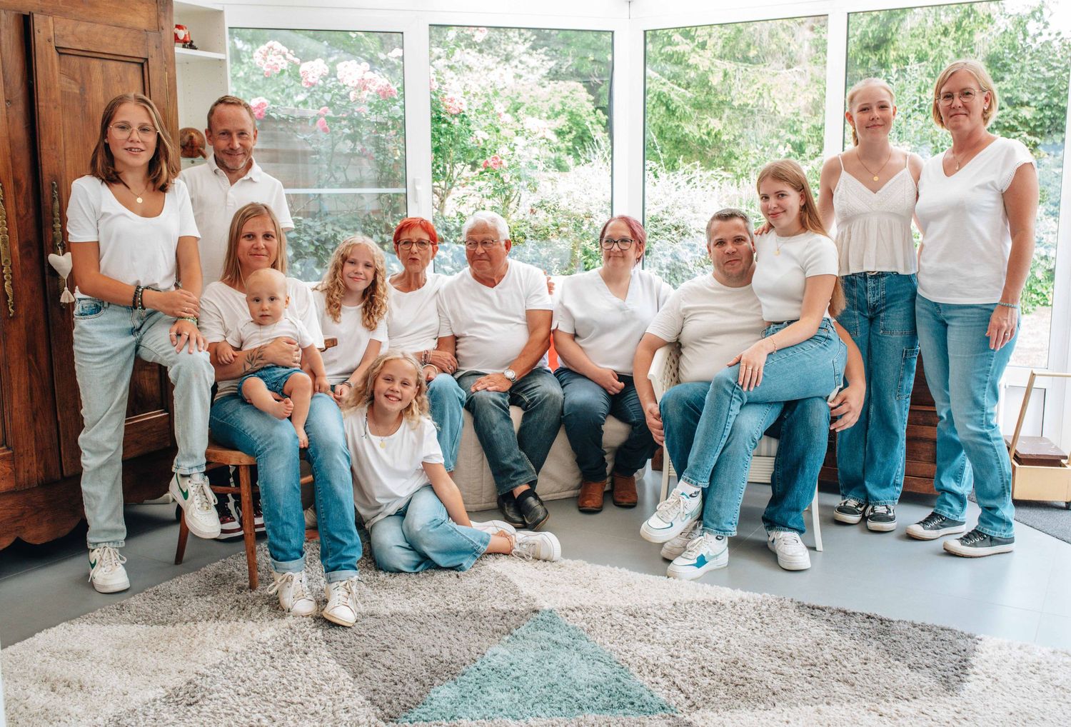 A large family group poses in coordinated white tops and blue jeans in a sunlit indoor space.