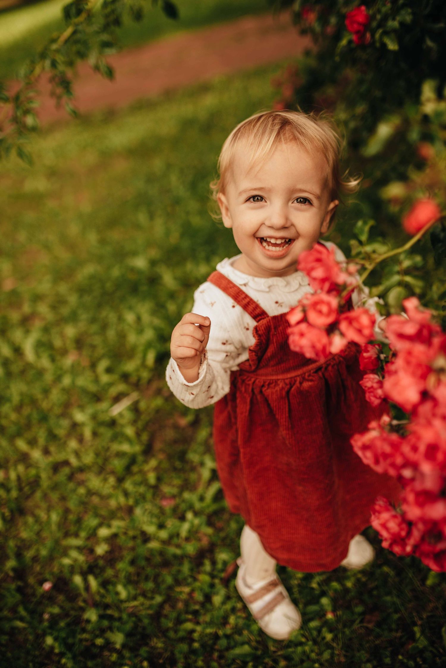 Toddler in red corduroy dress playing outdoors near pink flowers.