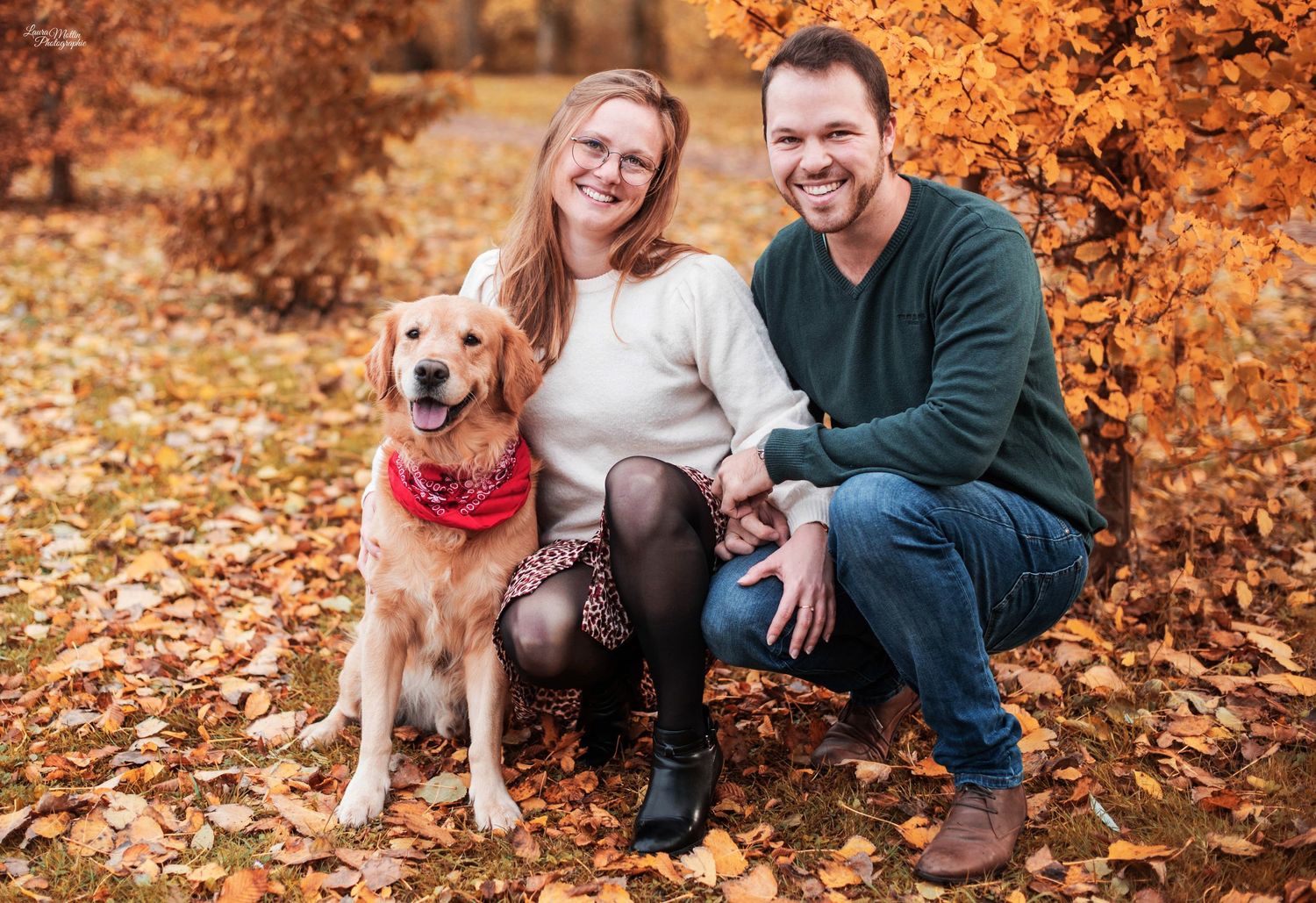 Happy couple sitting with their dog among fallen autumn leaves in a park setting.