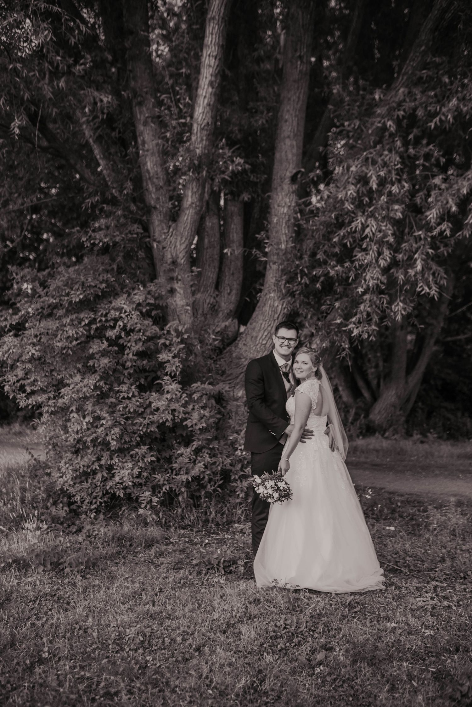 A black and white portrait of a couple in formal attire posing in a wooded setting.
