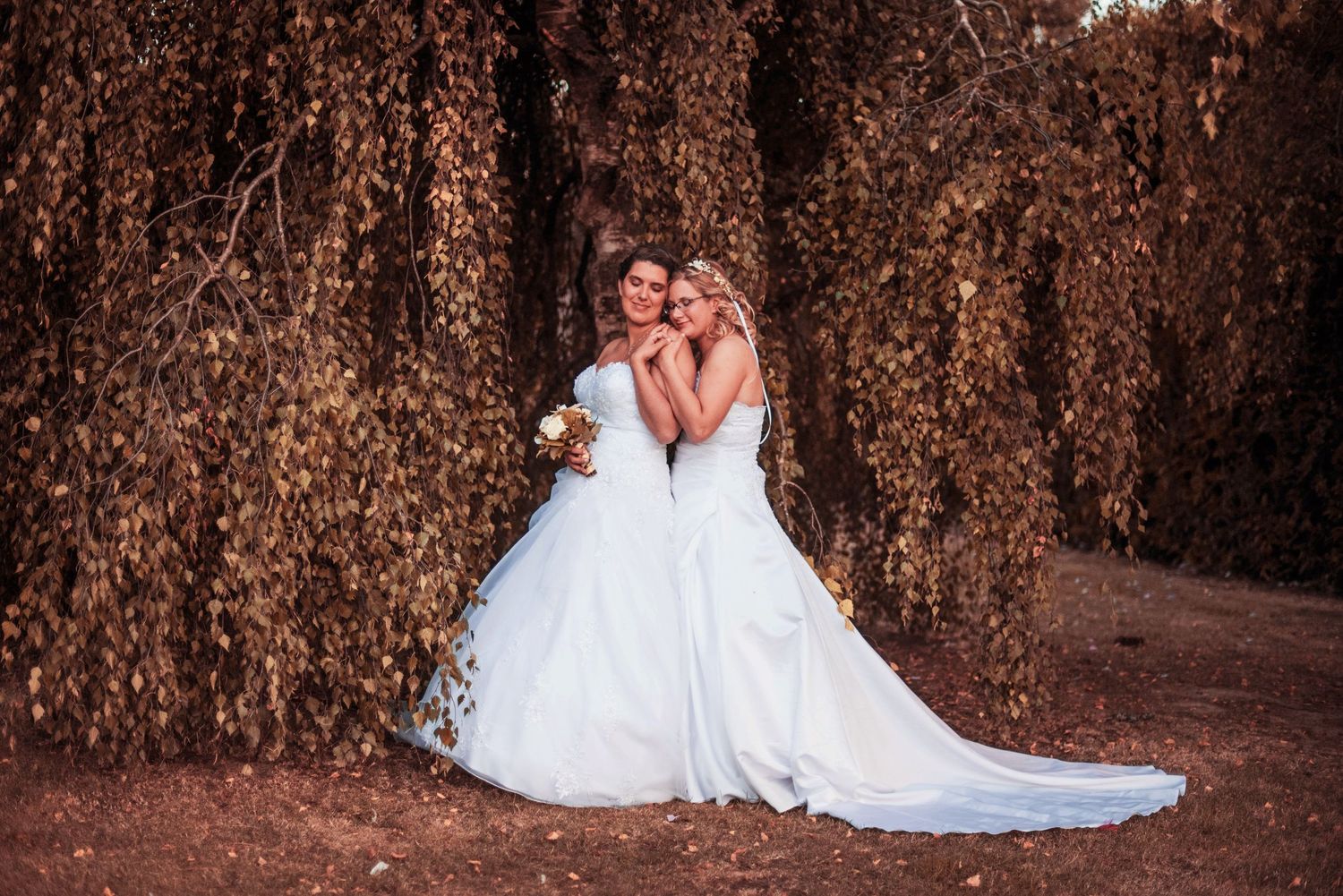 Two brides in flowing white gowns embrace against a backdrop of autumn foliage.