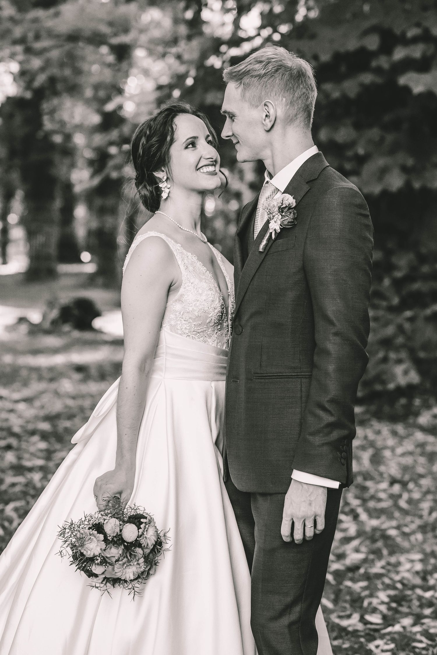 Black and white photo of a smiling bride holding a bouquet gazing at her groom in a suit outdoors.