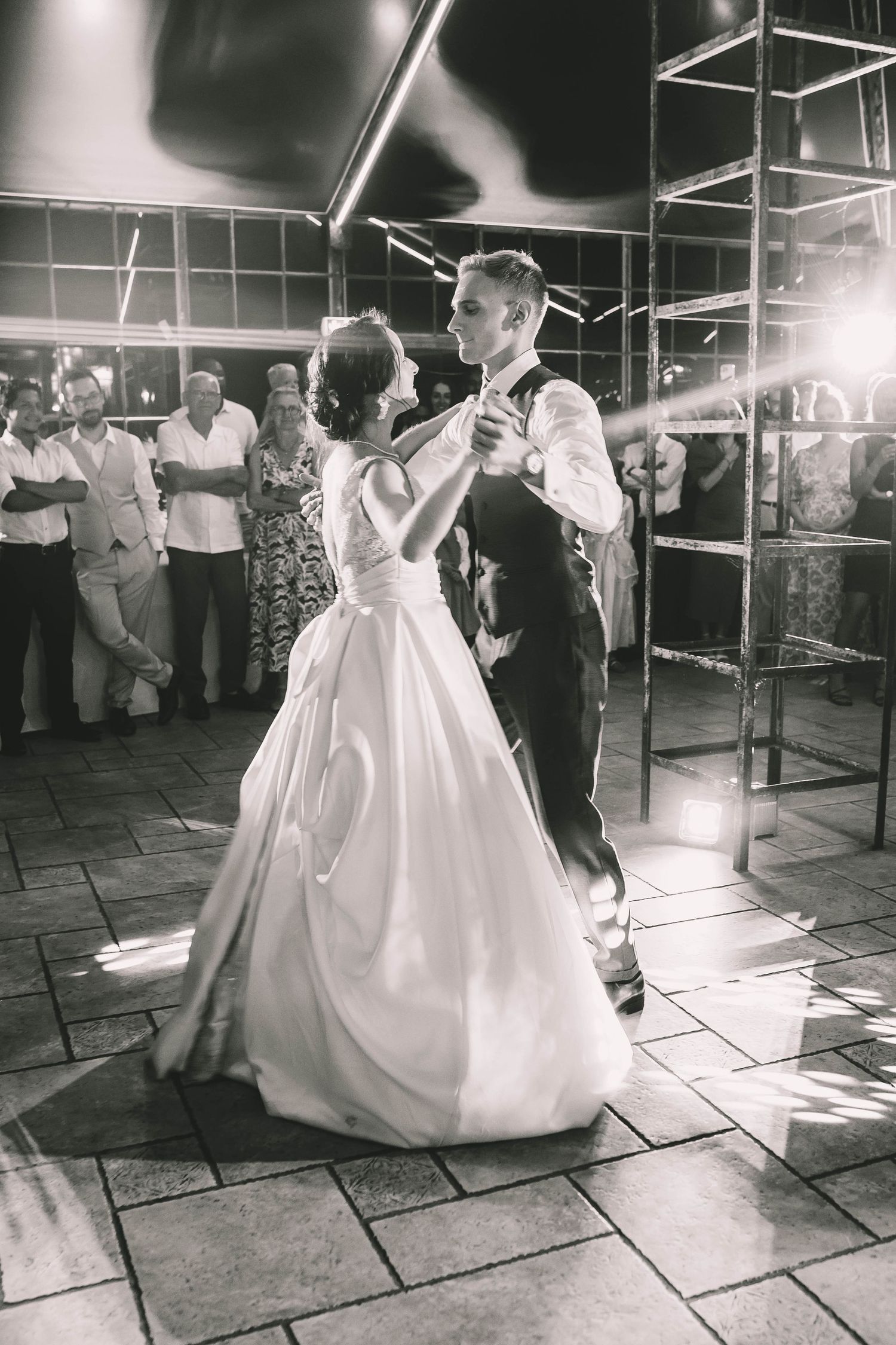 Black and white photo of bride and groom sharing their first dance at wedding reception with guests watching.