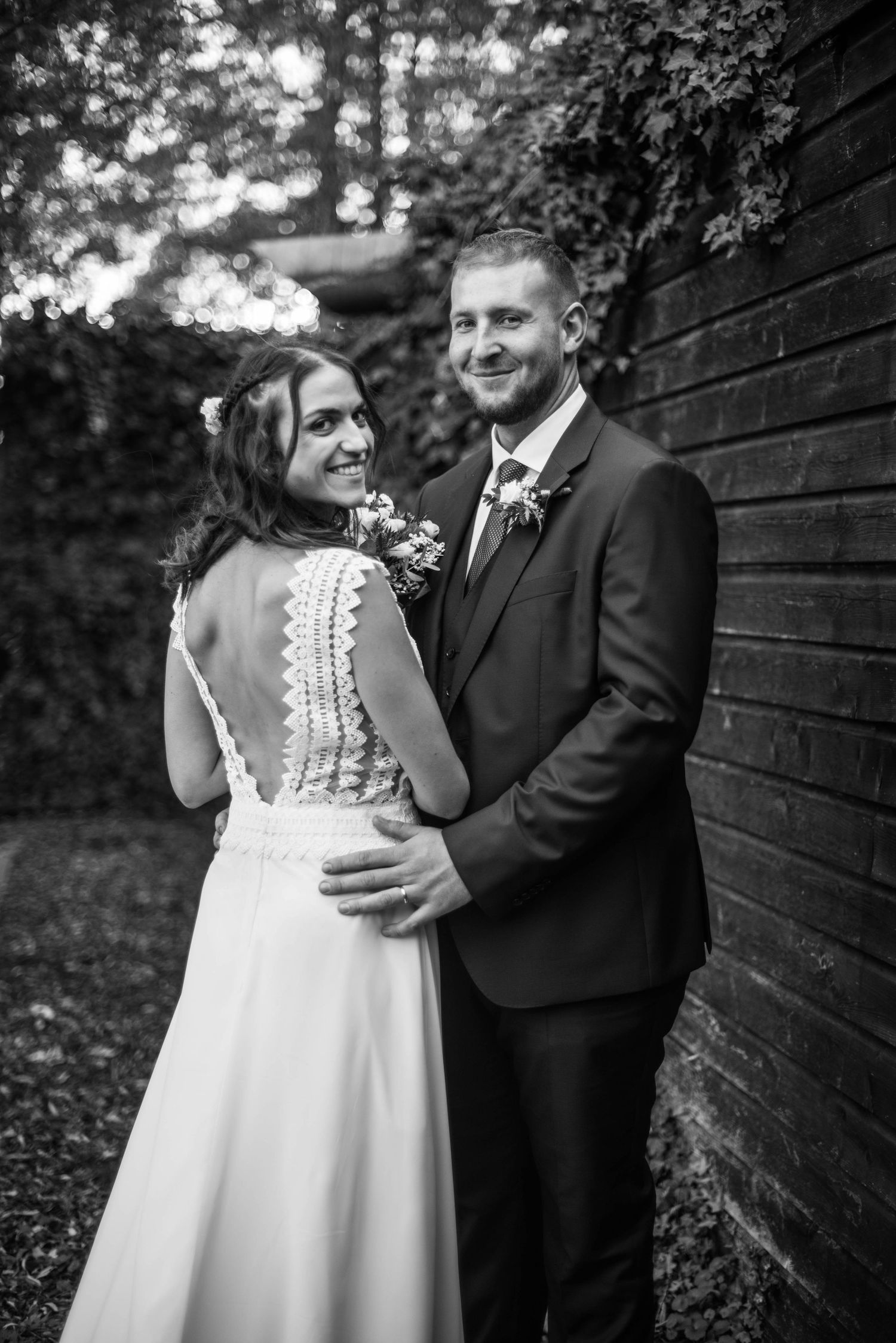 A black and white portrait of newlyweds posing against a rustic wooden wall outdoors.