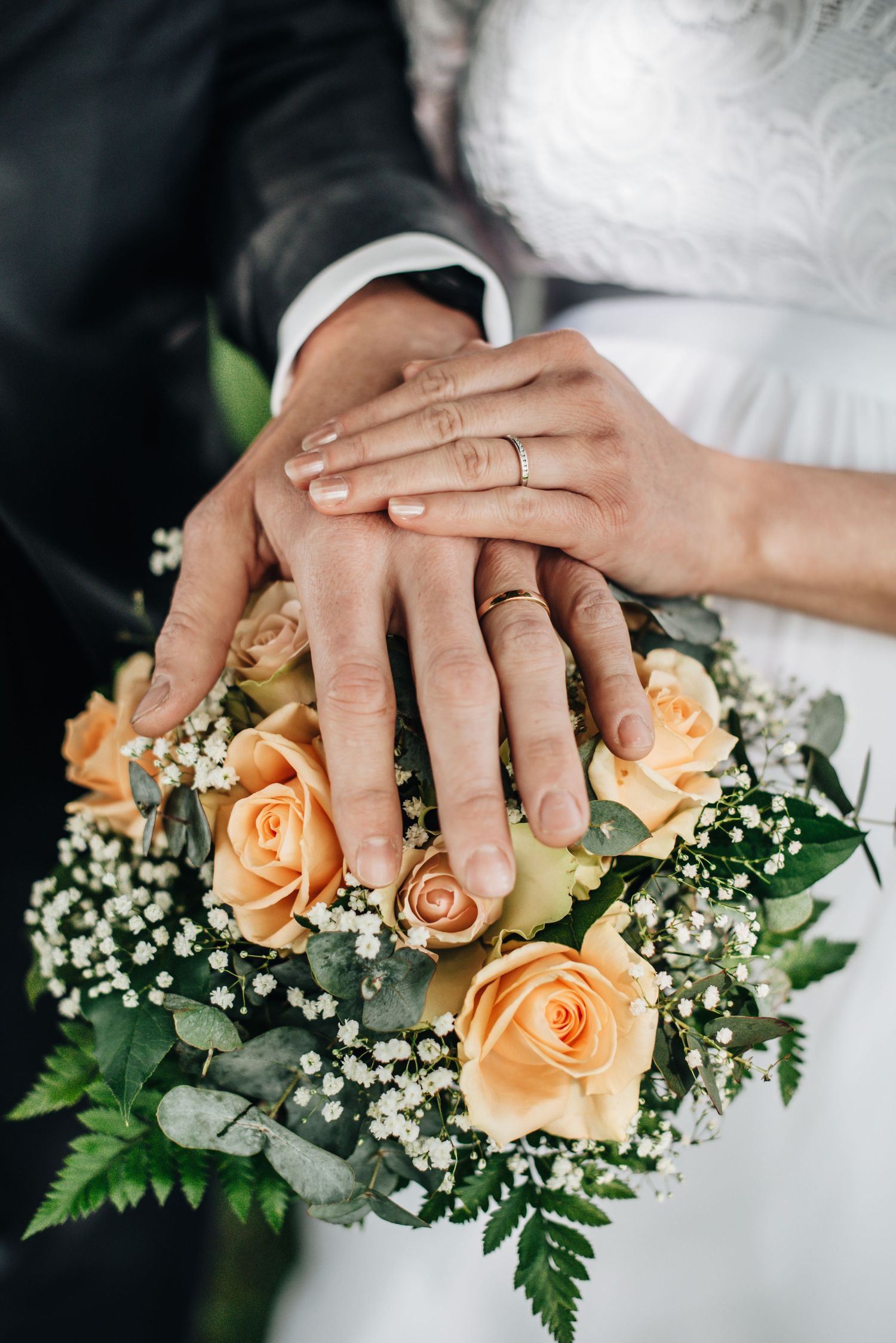 Close-up of hands with wedding rings resting on peach roses and baby's breath bouquet.