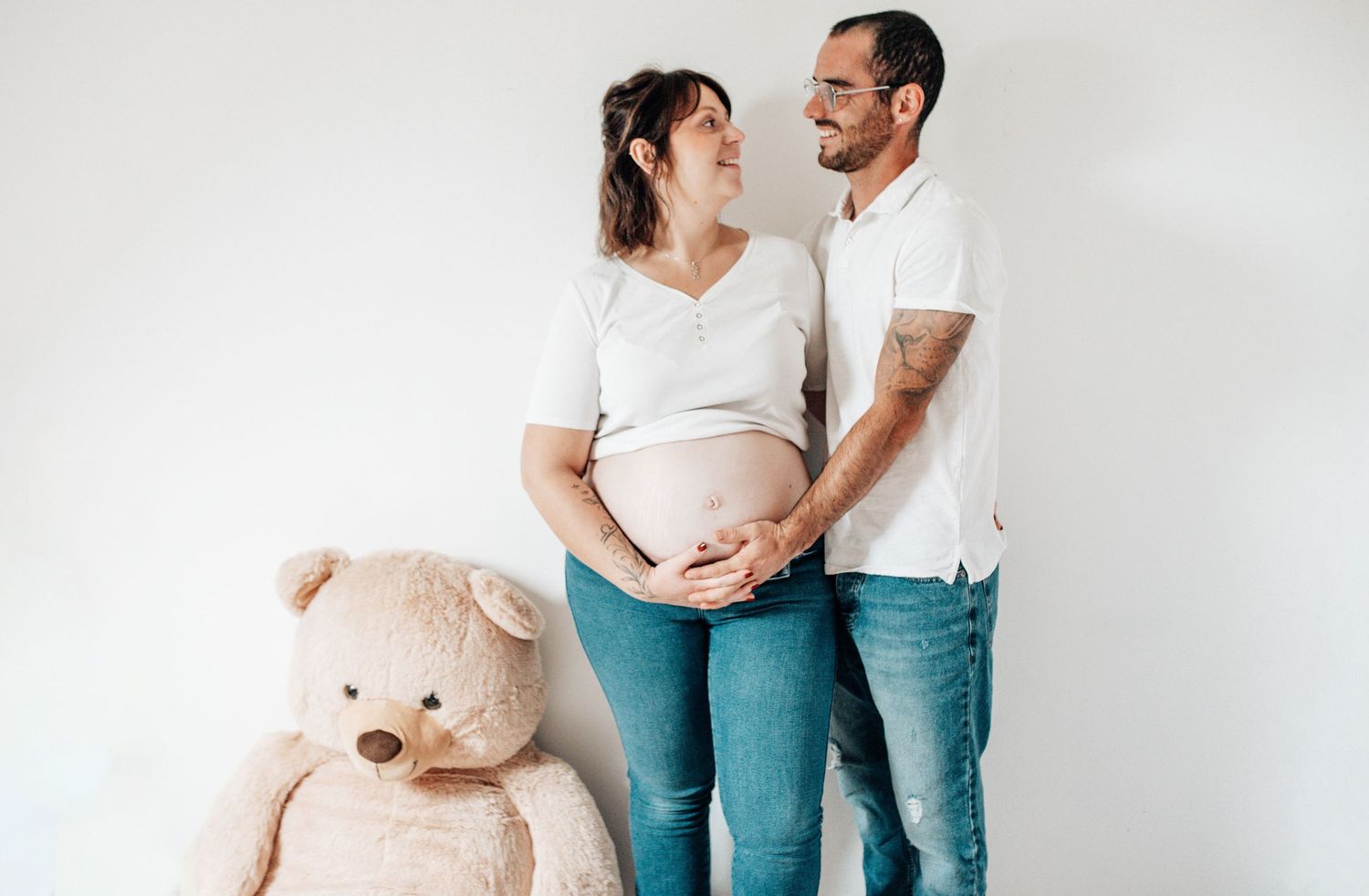 An expecting couple poses with a teddy bear against a white wall during a maternity photoshoot.
