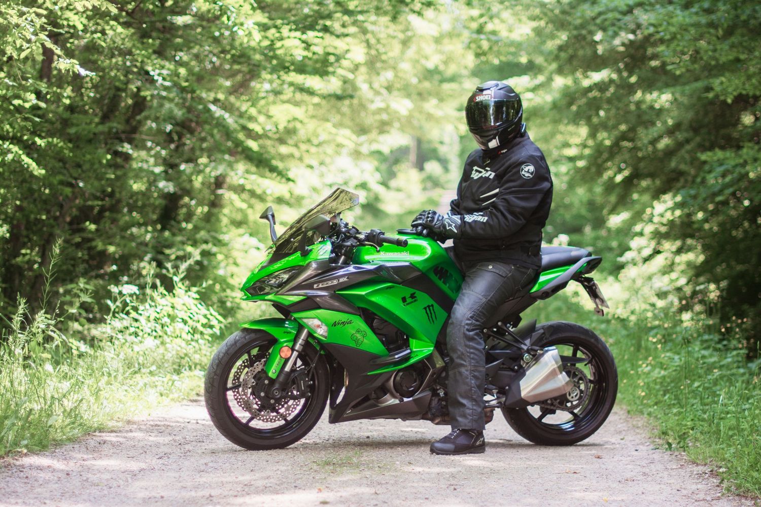 Rider in black gear stands with bright green Kawasaki motorcycle on forest path.