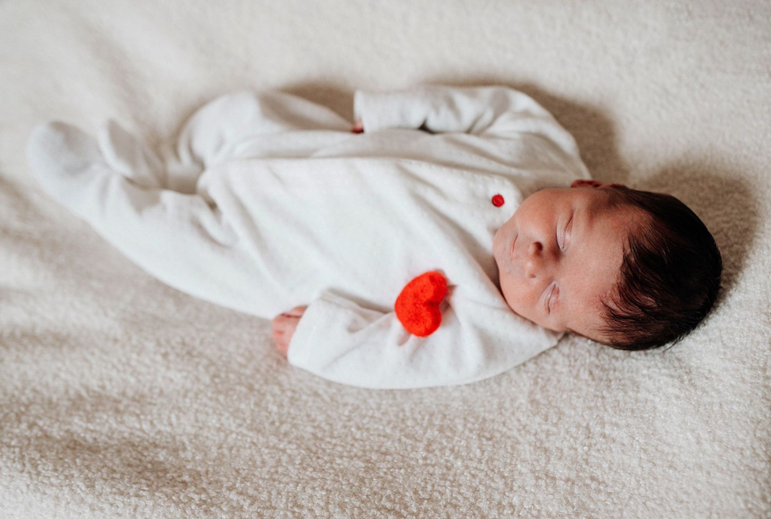 Newborn baby in white sleeper with red accent lies on soft white blanket.
