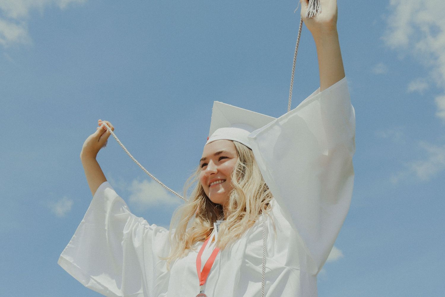 Image of graduate celebrating with arms up holding chords of honor while wearing cap and gown during photo shoot