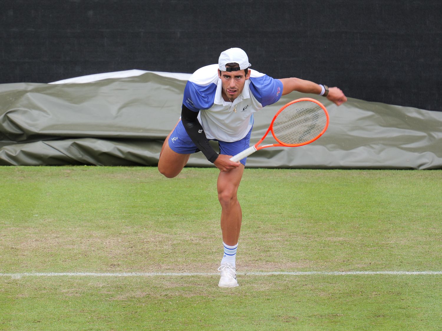 Tennis player serving on grass court, leaning forward with orange racket during a competitive match.