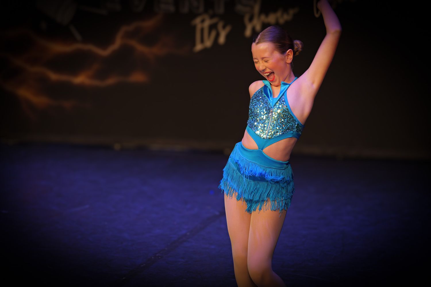 Young girl performing on stage in a sparkling turquoise dance costume with fringe skirt under colorful stage lights.