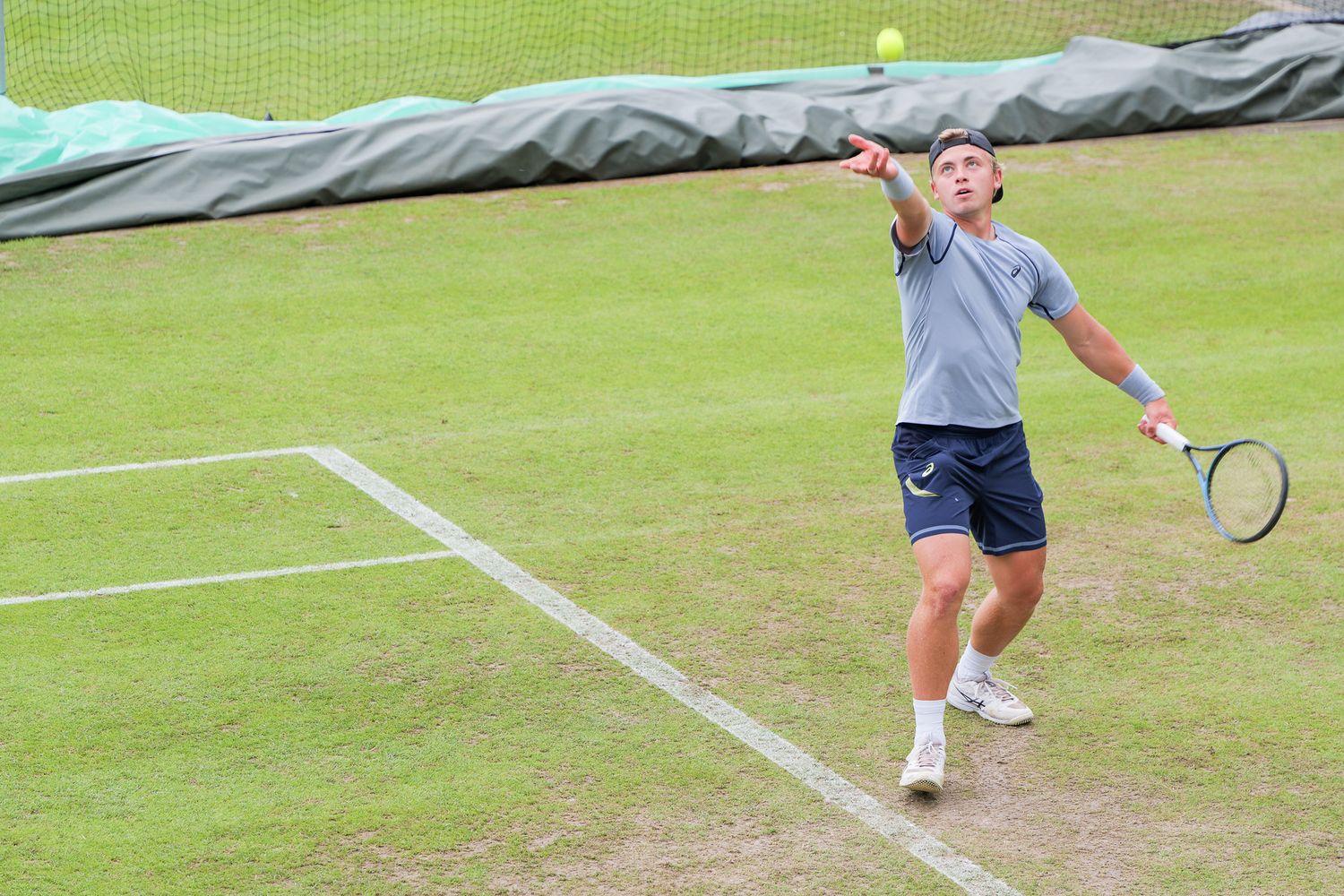 Male tennis player serving on a grass court, tossing ball overhead with racket raised during a competitive match.