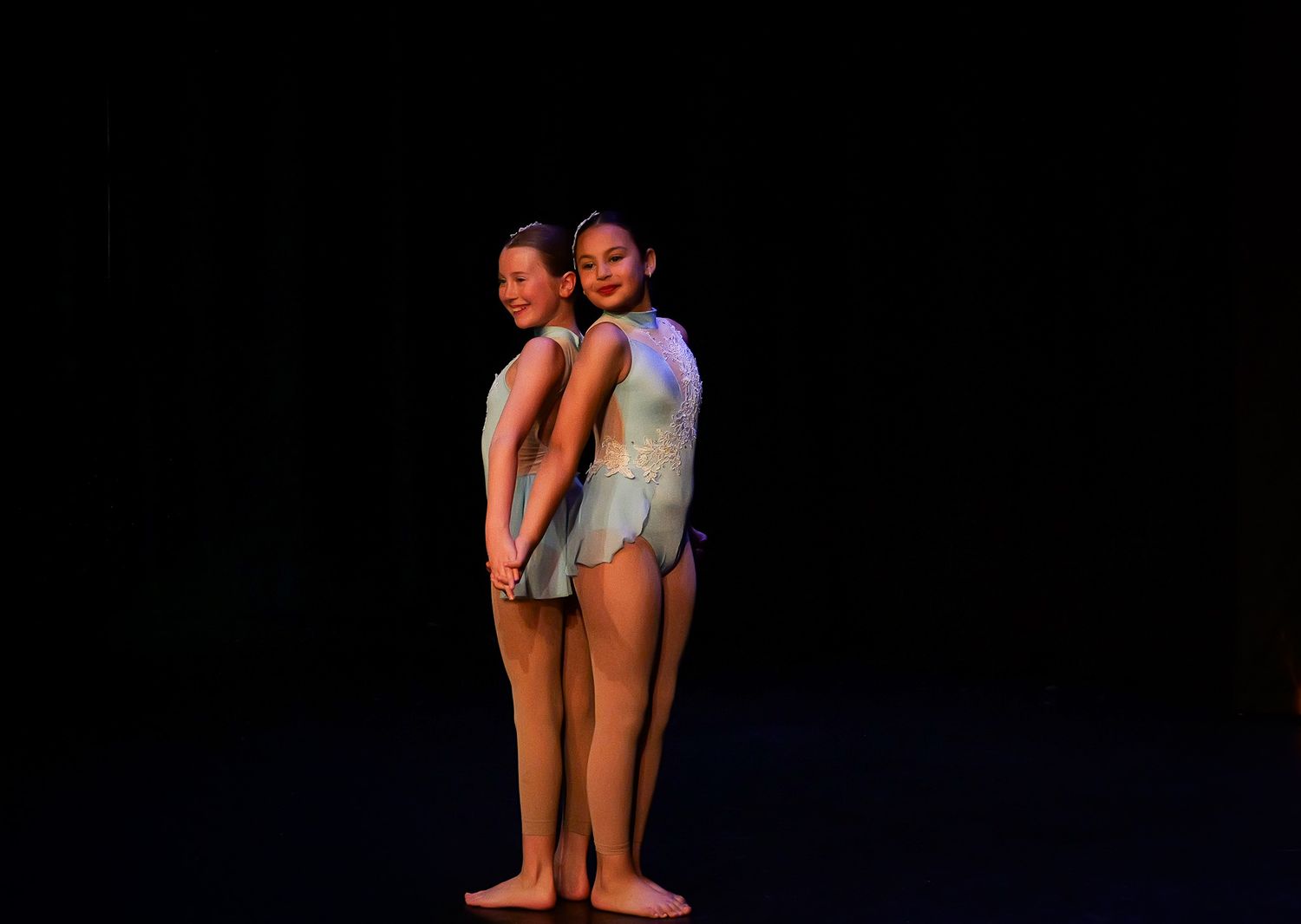 Two young dancers in leotards pose back-to-back on a dark stage during a dance recital performance.