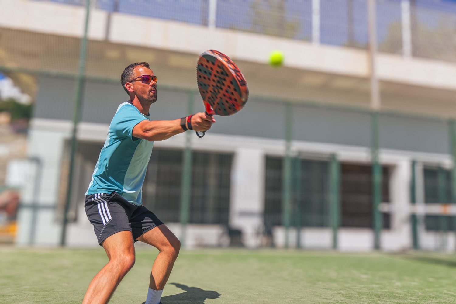 Man playing padel tennis, swinging a red paddle racket to hit a ball on an outdoor green court.