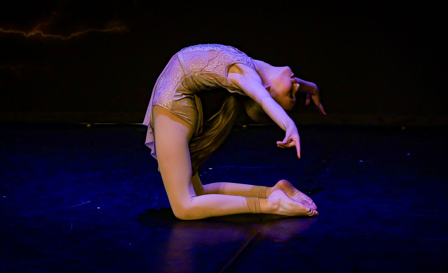A flexible dancer performs a deep backbend on a dark stage, wearing a beige costume under blue lighting.