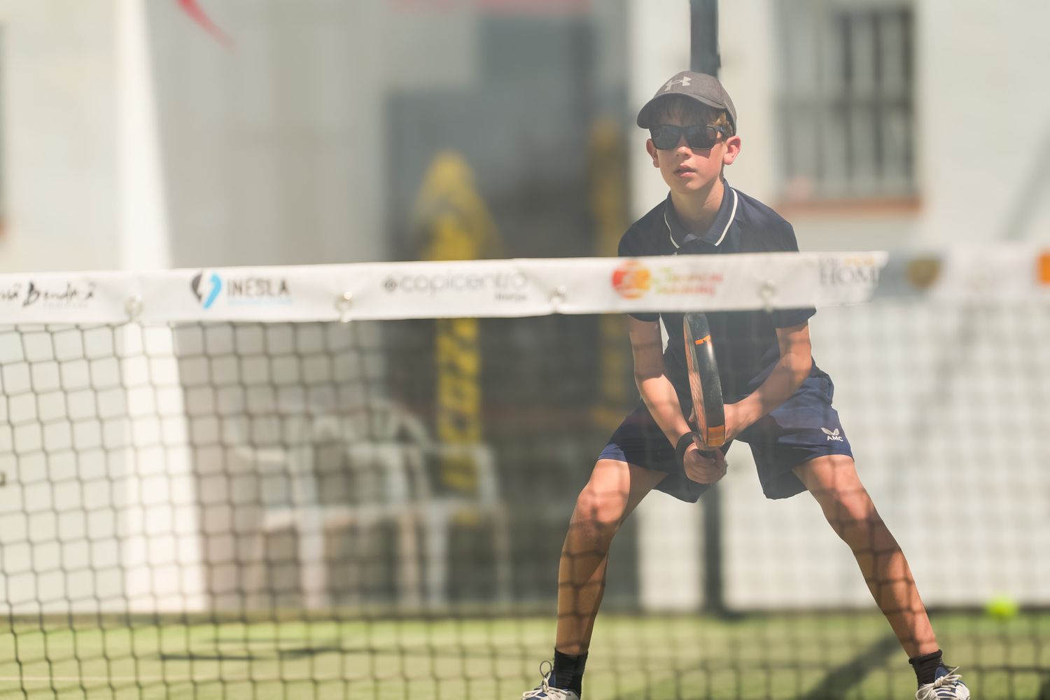 Young boy playing padel tennis near the net on an outdoor court, wearing a cap and sunglasses.