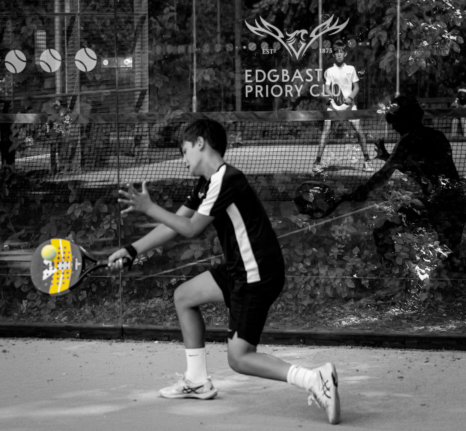Young boy playing padel tennis at Edgbaston Priory Club, lunging to hit ball with racket in black and white photo.