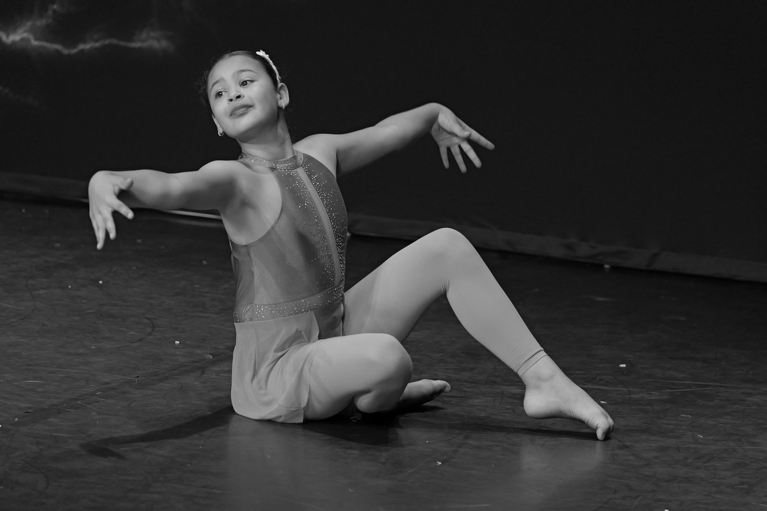 Young girl dancer poses gracefully on stage floor in leotard, arms extended, in black and white performance photo.