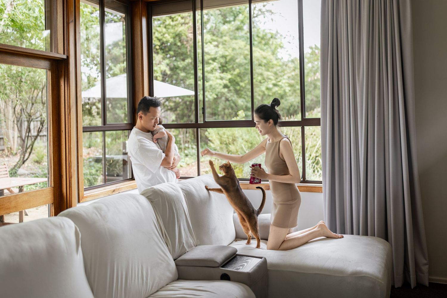 Couple relaxing on a white sofa near large windows with lush garden views indoors.